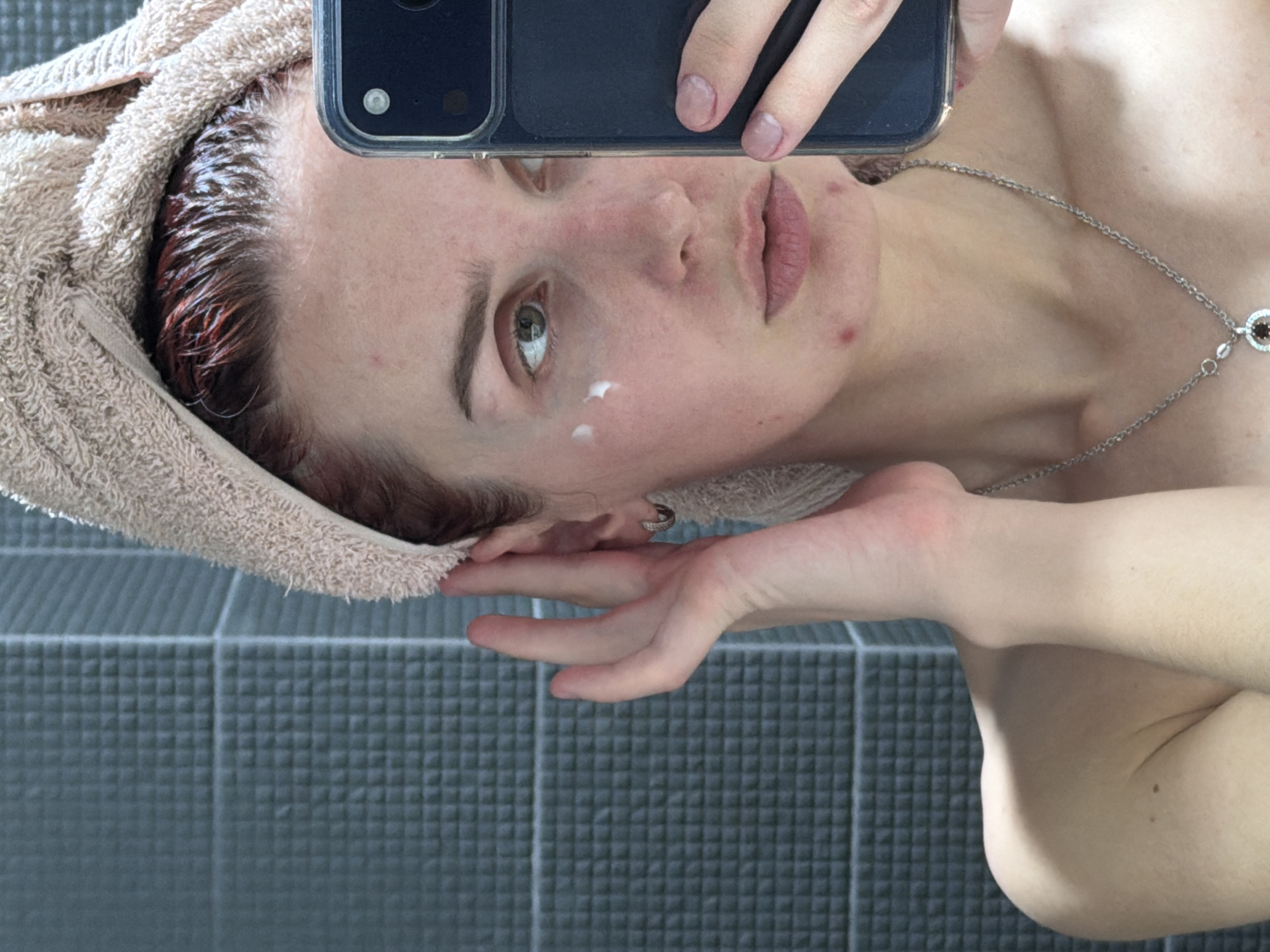 A person takes a selfie in a bathroom, likely in an apartment or house, in Chaikovsky, Russia. The individual has wet, reddish-brown hair wrapped in a beige towel. A smartphone is held up to the face, partially obscuring the view. There is a small amount of white cream applied to the left cheek. The person wears a silver necklace with a dark red pendant. The background features a wall tiled with small, dark grey square tiles.