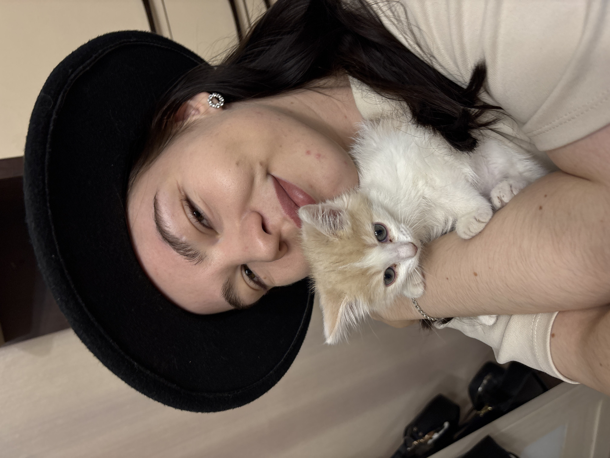 The image, taken in Perm, Russia, features a young adult woman cuddling a small kitten in an indoor setting. The woman is positioned on the left side of the frame, looking towards the camera with a slight smile or smirk. She has dark hair, wears a black wide-brimmed hat, and a light-colored top, with a circular silver earring visible in her left ear. A silver chain bracelet adorns her right wrist, which is seen holding the kitten.
The kitten, a small, fluffy creature, is predominantly white with light orange or cream patches, particularly on its head and ears. It has striking light blue or grey eyes and appears to be comfortably nestled in the woman's arms, also looking towards the camera.
The background is simple and out of focus, suggesting an indoor environment, likely a home. The walls appear to be light-colored. In the lower right background, some dark, out-of-focus objects, possibly bags or shoes on a shelf, are faintly visible. The lighting is soft, indicative of an indoor setting with ambient light. The overall mood conveyed is affectionate and warm, highlighting the bond between the woman and her pet. There is no visible text in the image.