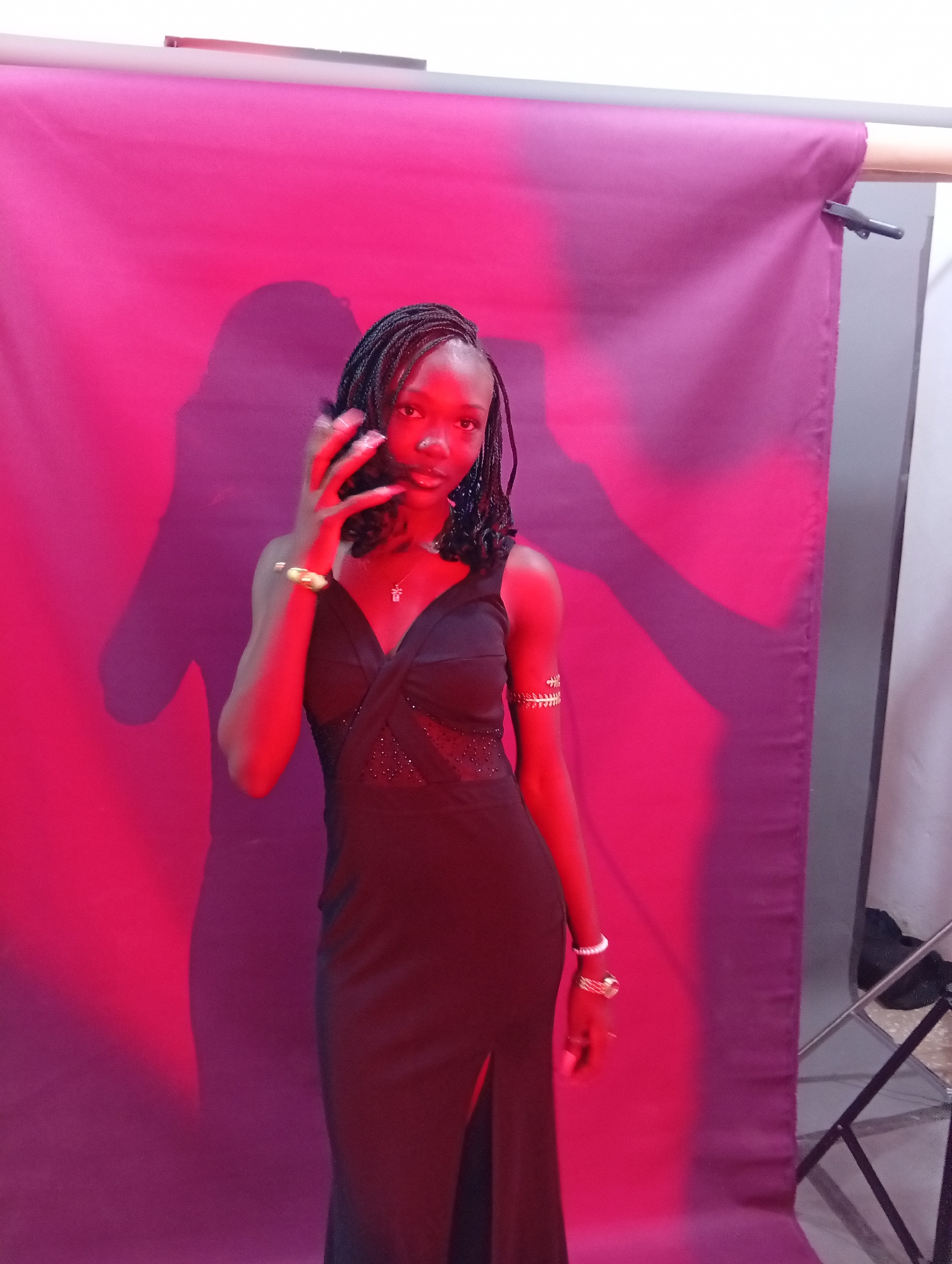 A young Black woman with braided hair stands in front of a purple backdrop illuminated by pink lighting. She is wearing a black, floor-length dress with a slit up the side and a v-neckline that features sheer fabric with embellishments. She has a gold bracelet on her right arm and several rings and a white bracelet on her left hand. Her left hand is raised near her face, with her fingers spread. There are shadows on the backdrop behind her, suggesting she is standing near a light source. A metal stand is visible on the right side of the frame. The image appears to have been taken indoors in a studio setting.