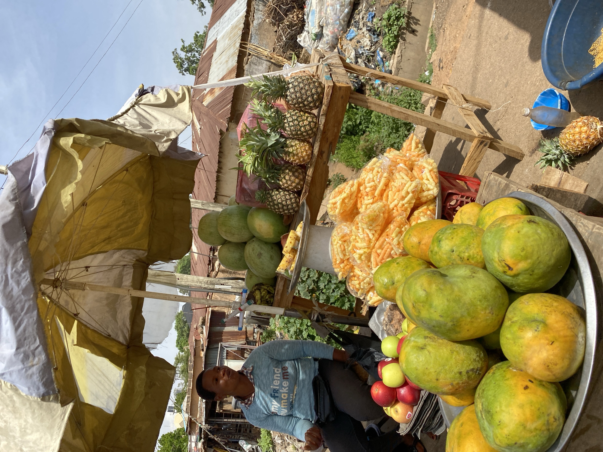 A vendor sits behind a makeshift fruit stand on a street in Bukuru, Nigeria. He is a Black man wearing a light blue long-sleeved shirt. The stand is laden with pineapples, melons, packaged orange snacks, papayas, and apples, displayed under a large yellow and white umbrella. The stand is constructed of rough-hewn wooden planks. In the background, there is a corrugated iron roof and plastic waste.