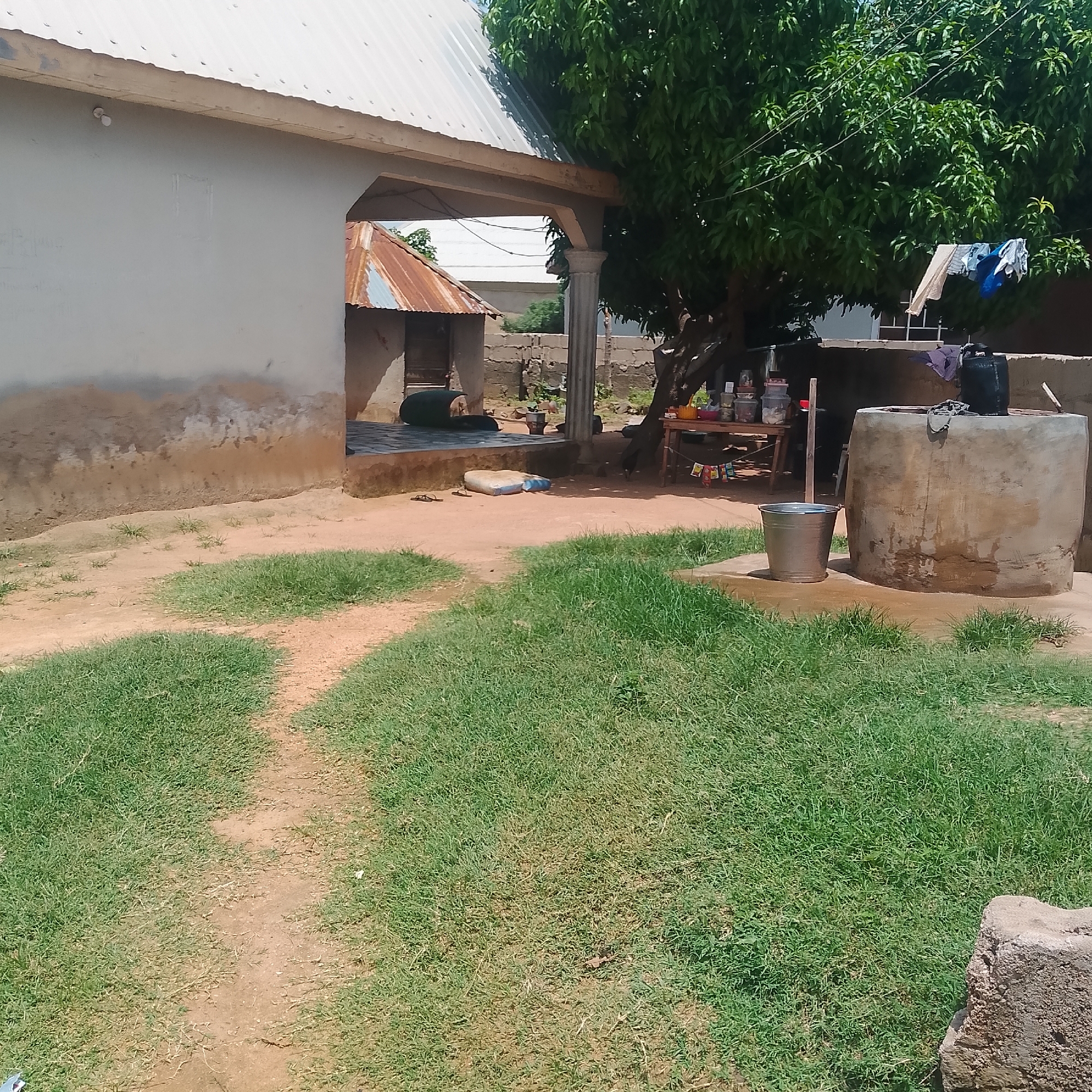 The image depicts an outdoor scene in Jalingo, Nigeria. The foreground features a dirt path winding through a patch of green grass. In the mid-ground, a light-colored, weathered building with a corrugated metal roof stands to the left. Part of another small building with a rusty metal roof is visible to the right of the main building, under an overhang supported by a column. Adjacent to this structure is a large, cylindrical concrete water well, with a black container on top. To the left of the well, a metallic bucket sits on the ground.
A large tree with dense green foliage is prominent in the background, casting shade. Beneath the tree, a wooden table holds various containers and items, including what appear to be jars or bottles. Laundry, including a blue garment and a light-colored fabric, is hanging on a clothesline strung between the tree and another point. The ground is a mix of dirt and sand. The overall impression is of a residential or communal area, possibly a compound, with elements of daily life such as water storage and laundry visible. The bright lighting suggests it is daytime, likely sunny. There are no visible people or text in the image.