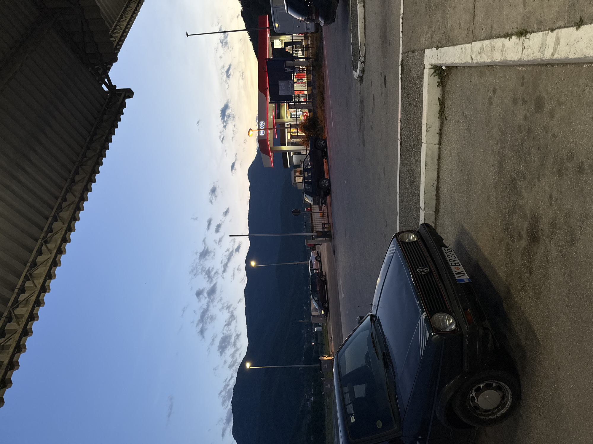 The image depicts a street scene in Mojkovac, Montenegro, at twilight. An older model, dark-colored Volkswagen car is parked on the right side of the frame, partially obscuring the foreground. Its license plate reads "NK BR378."
In the background, across the street, is an EKO gas station with its red and white signage clearly visible. Several cars are parked near the gas station, indicating activity. A mountain range forms a backdrop to the town and gas station, partially obscured by the descending twilight sky.
Streetlights line the road, illuminating the scene. The left side of the frame is dominated by the underside of a metal roof structure, presumably belonging to a building or shelter. The road appears to be paved and lined with a concrete sidewalk, which is visible on the right side of the frame.