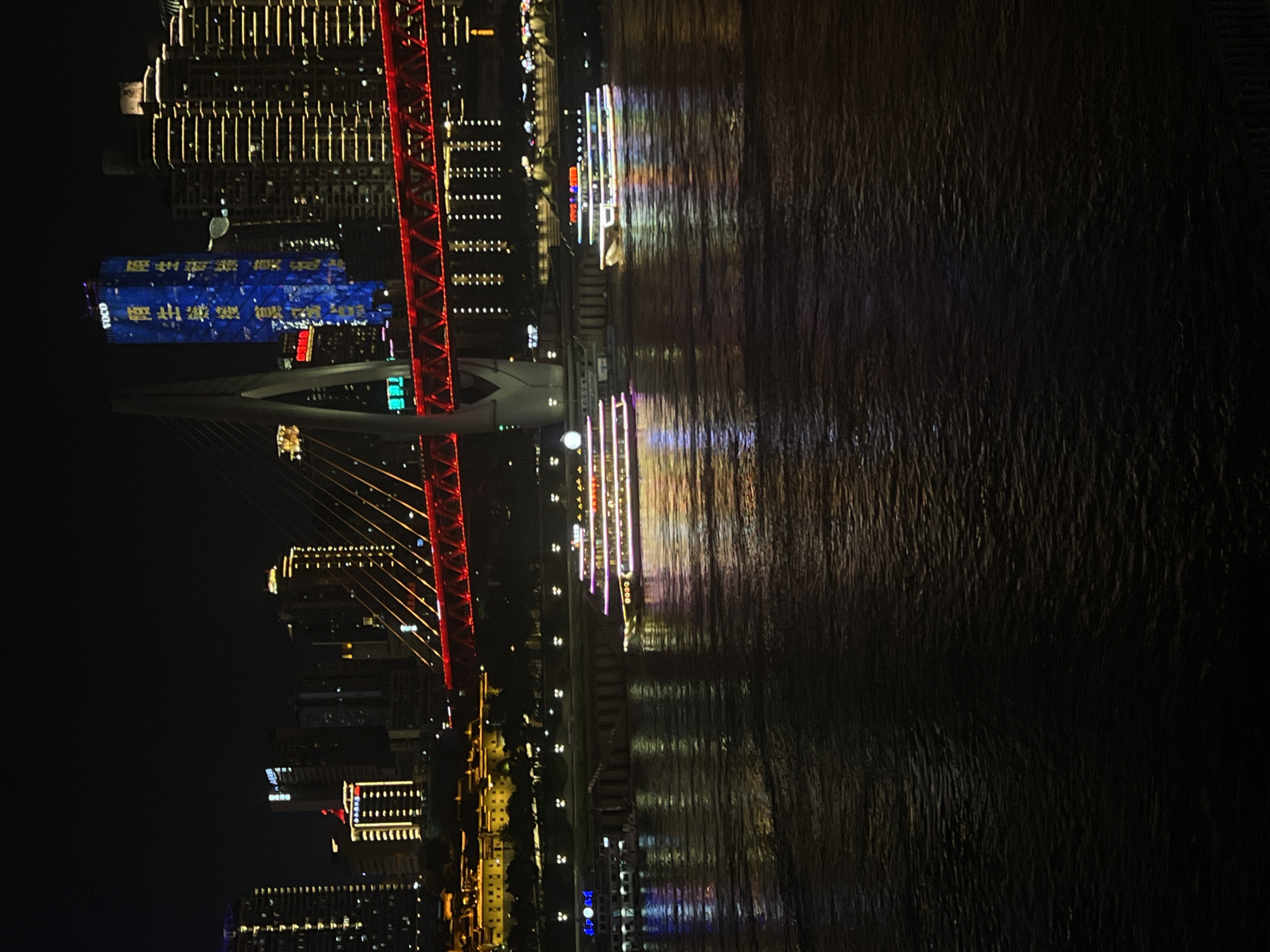 The image depicts a night view of the Yangtze River in the Nan'an District of Chongqing, China. A large, modern bridge with red illuminated accents spans the river, connecting the Nan'an District to the Yuzhong District. The bridge's concrete support structure is visible on the left. Two illuminated tour boats are navigating the dark, reflective waters of the river. The city skyline on both sides of the river is adorned with numerous bright lights from skyscrapers and buildings. One prominent blue skyscraper features illuminated Chinese characters and the logo "VOCO". The overall scene captures the vibrant nightlife and urban landscape of Chongqing.