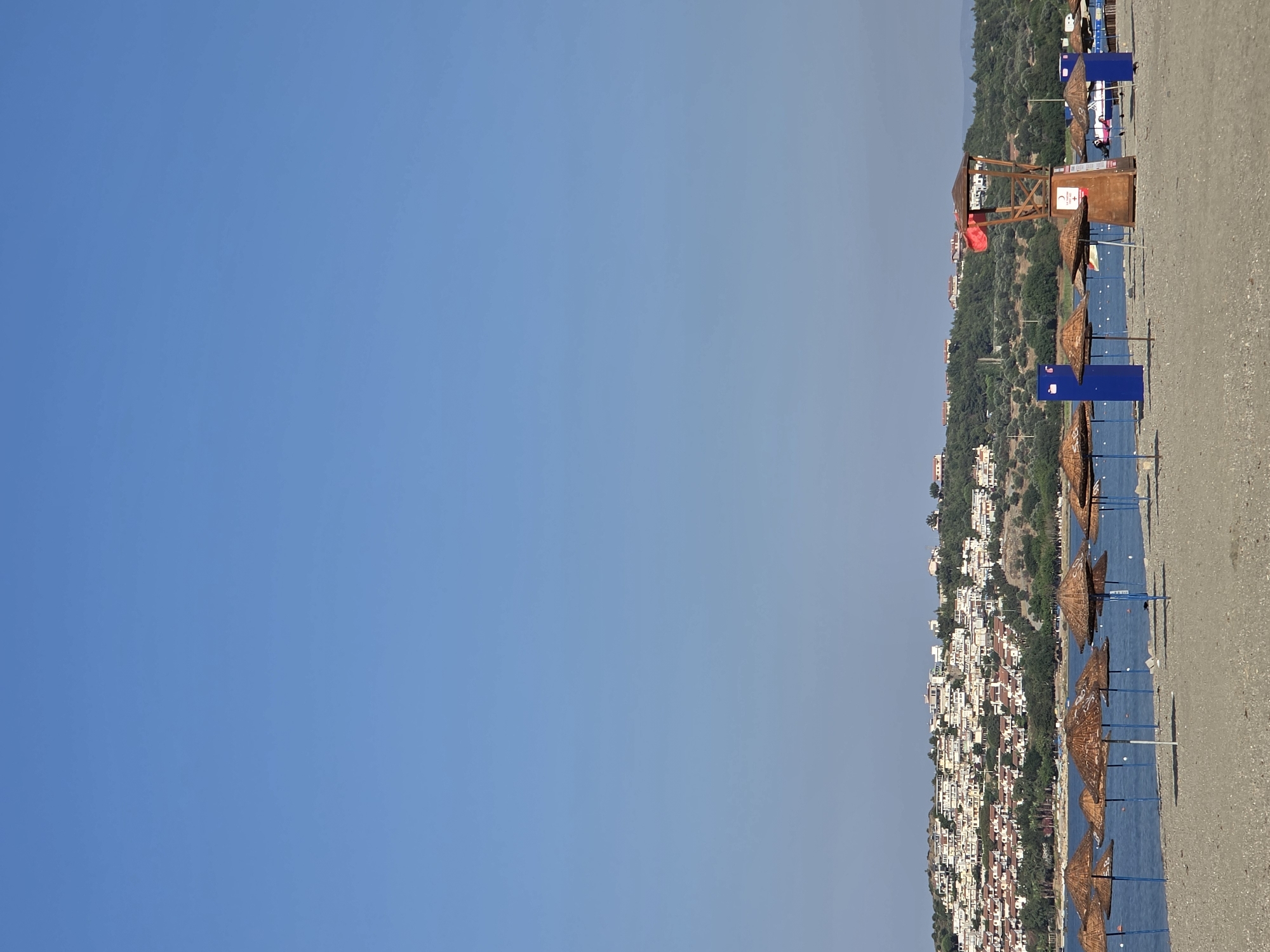 The image shows a beach scene in Seferihisar, Turkey. In the foreground, a sandy beach is lined with straw umbrellas and a lifeguard tower. The lifeguard tower is made of wood and has a red flag flying. Behind the beach, the blue sea is visible, with a hillside covered in white houses and green trees in the background. The sky is clear and blue.