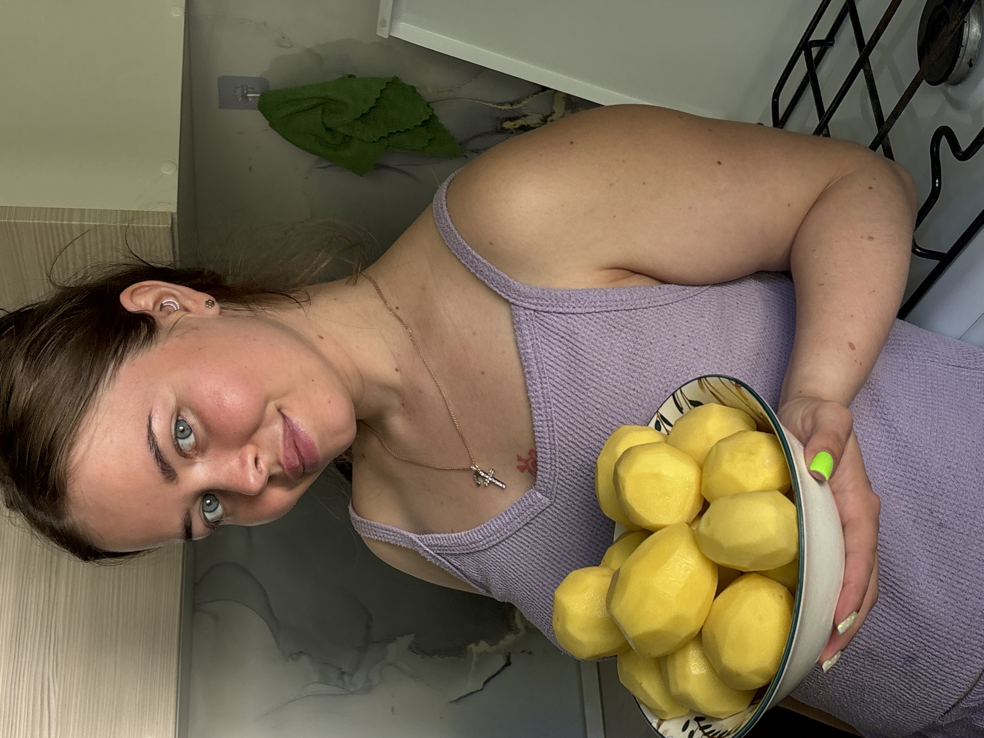 A fair-skinned woman with brown hair and blue eyes is holding a bowl of peeled potatoes. She is wearing a lilac tank top and a gold necklace with a cross pendant. Her nails are painted, with one hand having a neon green accent nail. There is a green cleaning cloth visible on a kitchen shelf in the background, along with a portion of a gas stove. The location in the city of Izhevsk, Russia, cannot be determined from the image.