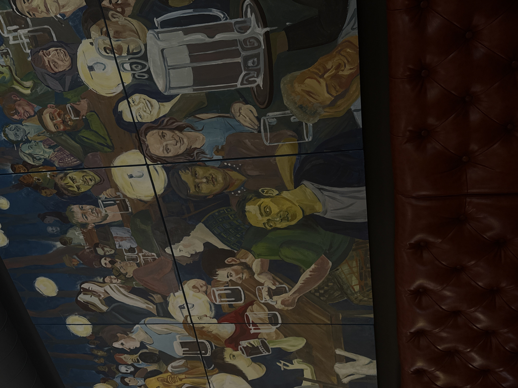 The image is a close-up of a ceiling mural in a building located in Perm, Russia. The mural depicts a crowd of people gathered around tables and holding beer mugs. The style of the mural is illustrative, with some figures simplified or stylized. The scene appears to be a lively bar or pub atmosphere. The mural is composed of individual panels, with visible seams between each. To the right of the mural, a tufted leather-like panel is seen, likely a wall decoration.