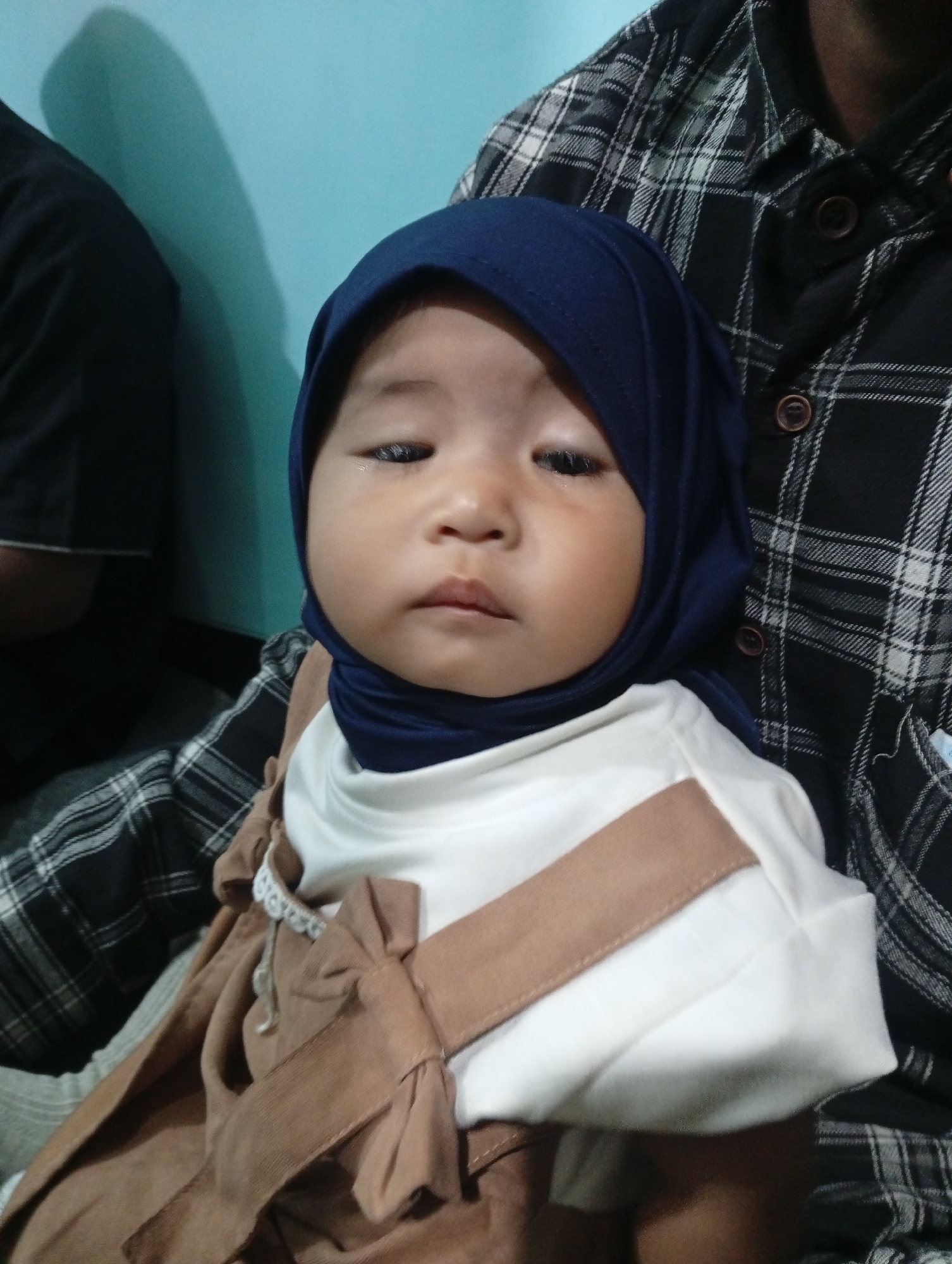 A close-up, waist-up shot shows a young child wearing a blue hijab and a brown dress over a white shirt. The child is held by an adult wearing a black and white plaid shirt. The background is a teal-colored wall. The scene appears to be indoors, likely a residential setting in Jatisari, Indonesia.