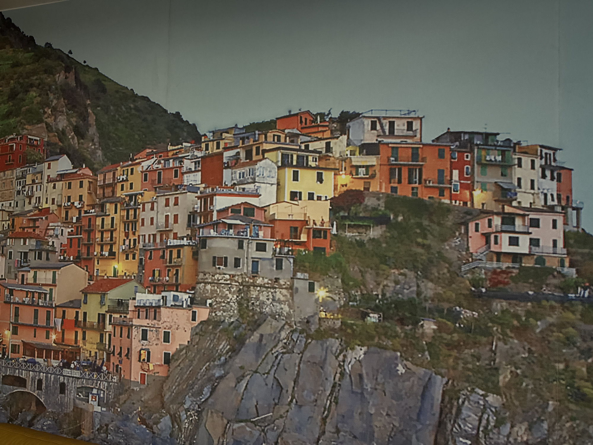 The image depicts a coastal village built on a steep hillside, likely in Italy, rather than Stara Zagora, Bulgaria. The village consists of closely packed, colorful buildings with terra cotta roofs and windows, clinging to the rocky terrain. A bridge with people on it is visible in the lower left, and the hillside is covered with vegetation. The sky is a muted gray. The overall impression is a scenic, Mediterranean vista.