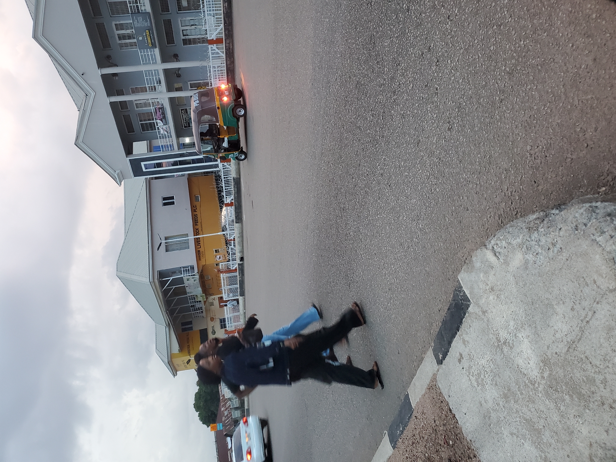 The image depicts a street scene in Jos, Nigeria. In the foreground, a concrete sidewalk with black and white markings lines the asphalt road. Three people are walking on the road, two of whom appear to be crossing. The person in the forefront is wearing a dark blue jacket and black pants, while the person behind them wears a black shirt and blue jeans. Further back, a white car is visible on the left, and a colorful auto-rickshaw, known locally as an "achaba," is moving on the right side of the road. The auto-rickshaw is green and brown and has "PIA 1035" written on its back. Behind the road, a modern building with multiple stories and balconies stands, featuring signs for "LIVESTOCK FEEDS PLC." and "Essence by GIMBAH". The sky is overcast with gray clouds.