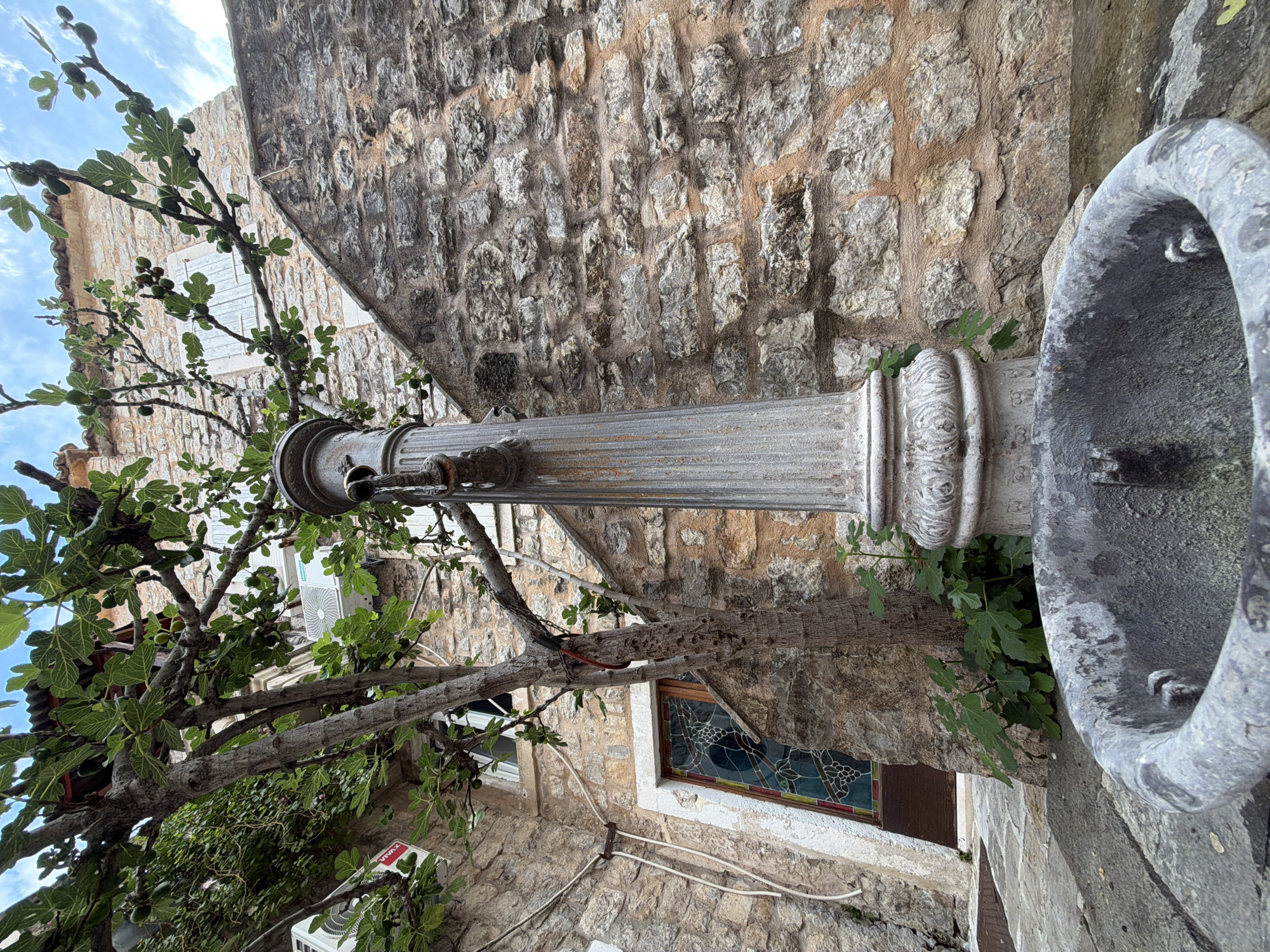 The image shows a public drinking fountain in Budva, Montenegro, with a stone wall behind it and a fig tree to the left. The fountain is made of metal and appears to be old. The stone wall is made of light-colored stones and has some vegetation growing on it. The fig tree has green leaves and figs. There are buildings visible in the background, and the sky is blue with some clouds.