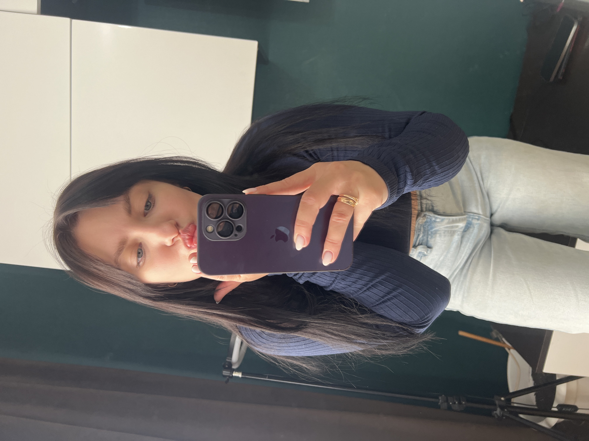 The image shows a young woman taking a mirror selfie indoors in what appears to be a studio or a room equipped for photography, located in Марковский, Russia. She has long, dark hair and light eyes, and is making a playful "kissy face" gesture towards the camera.
The woman is holding a dark purple iPhone with a visible Apple logo and multiple camera lenses in her left hand. A gold ring adorns her ring finger, and her nails are neatly manicured. She is wearing a dark blue ribbed long-sleeve top and light blue denim jeans.
The background consists of a bright white wall or cabinet on the left side of the frame, and a dark teal or green wall or backdrop on the right. Towards the bottom right, parts of black photography equipment, including a tripod, are visible, suggesting a controlled environment for shooting. The lighting appears bright and even, consistent with an indoor setting. The overall expression of the woman conveys a casual and lighthearted mood.