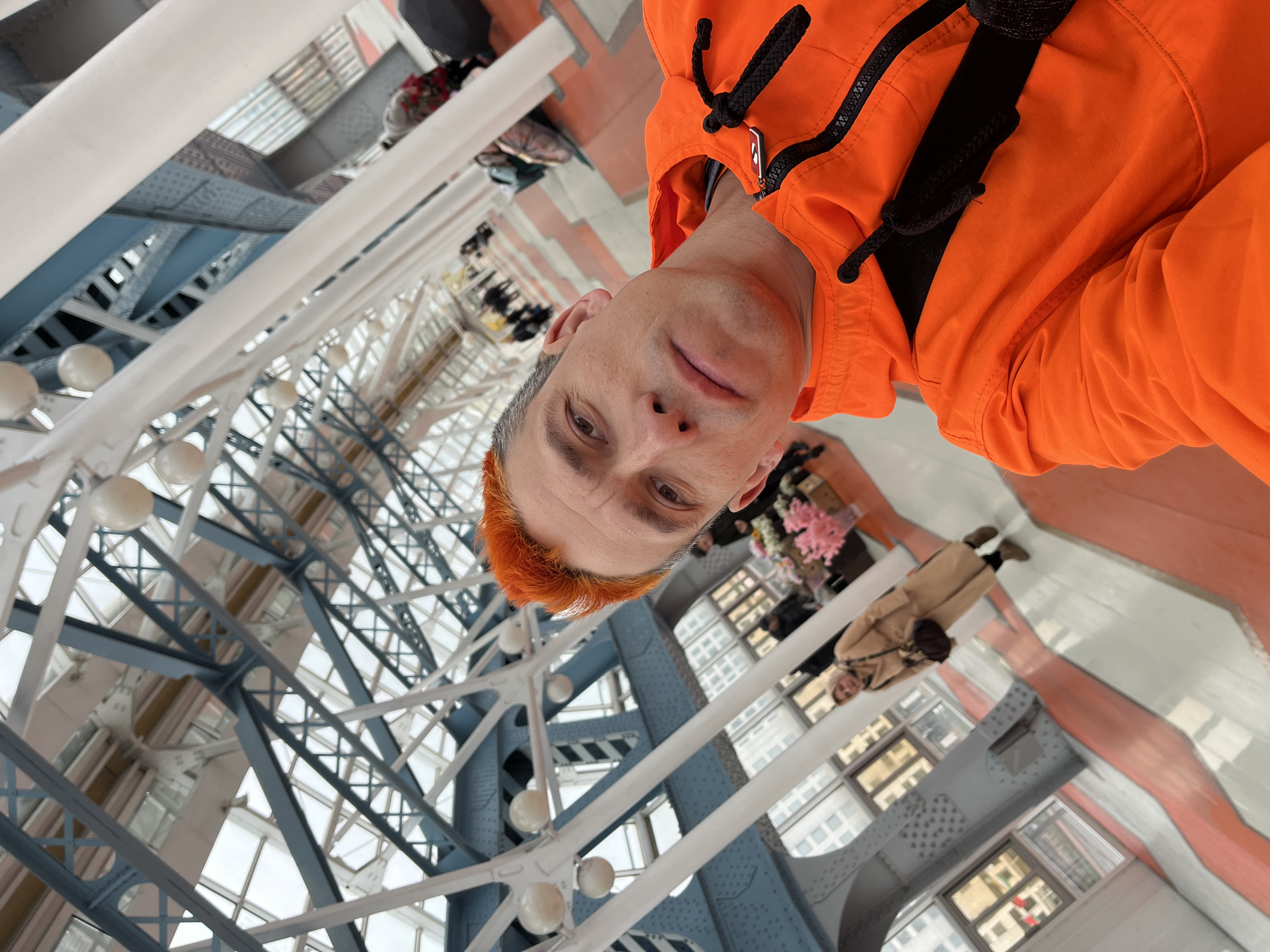 A man in an orange hoodie is taking a selfie. He has orange hair. The background includes a steel structure with windows, and people walking on an elevated platform. Based on the steel structure and architecture, the location is likely the glass-bottomed bridge at Zaryadye Park in Moscow, Russia.