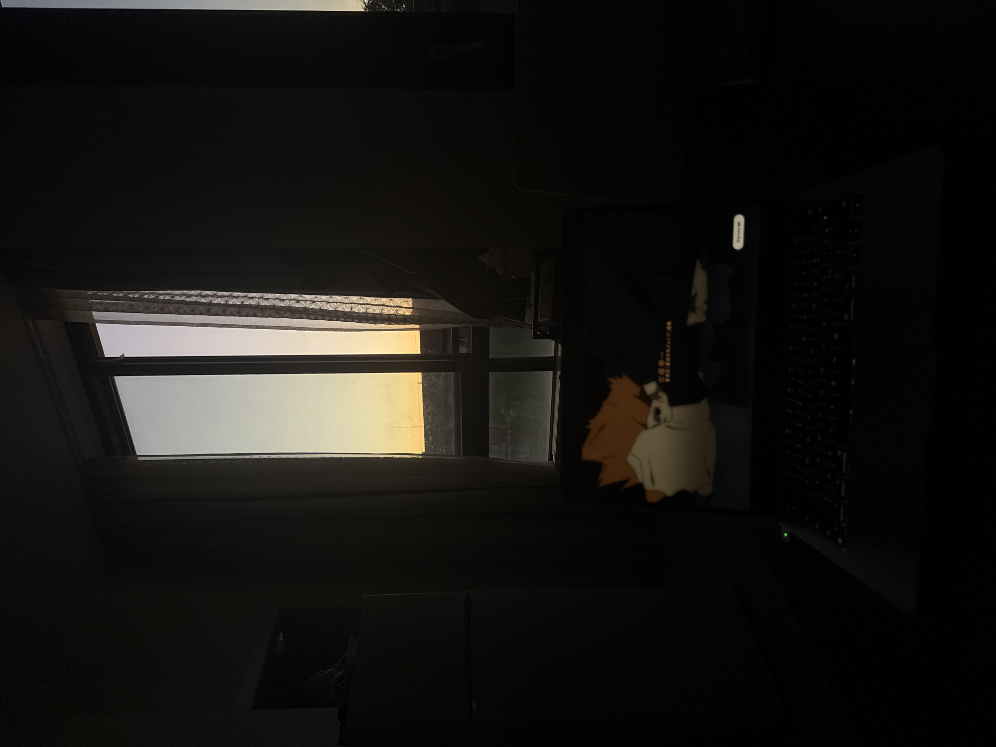 A laptop sits on a desk in a dark room. The laptop screen displays an image of an anime character with orange hair. A window is visible behind the laptop, showing a view of the sky with a yellow-orange gradient. The room appears to be indoors and the photograph is taken in the evening or early morning. The location cannot be determined to be Milan, Italy from the image.