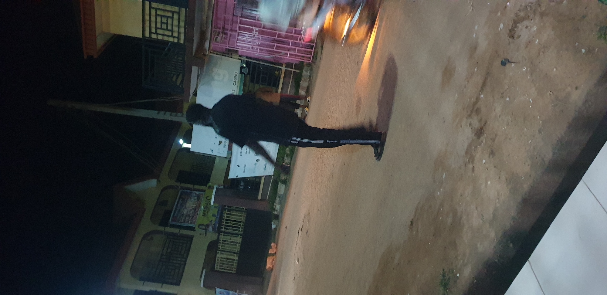 This image captures a nighttime street scene in Narayi, Nigeria, depicting an unpaved road flanked by multi-story buildings under artificial illumination.
In the foreground, a person is seen from behind, slightly blurred, indicating movement or a long exposure. This individual wears a black short-sleeved shirt and black track pants featuring three vertical white stripes on the side, with the word "Supreme" visible vertically on one of the stripes. Dark sandals are worn on their feet.
To the right of the primary subject, a blurred motorcycle or scooter is in motion, casting an orange light forward or from its tail light onto the road. Further in the mid-ground, a second person is partially visible, wearing lighter-colored footwear.
The left side of the scene features a two-story building, light yellow or beige with a reddish-brown roof trim. It has balconies with dark metal railings, one displaying a geometric pattern and another with vertical bars. A prominent banner on this building advertises "MAMA J C" alongside a woman's image. Below, a dark metal gate encloses the property.
On the right side of the frame, another building with a reddish-brown roof trim is partially visible, featuring dark metal railings with a distinct geometric design. A pink or purple metal gate or barrier stands on the far right. A tall, slender utility pole, likely concrete or wood, rises on the right side, supporting overhead wires.
Two white banners are displayed on posts or structures in the mid-ground. One banner has partially legible text including "INSTAN" and "ILLS CASINO." The other banner contains text such as "DISTRIBUT..." and "Clean Agi..."
The ground consists of an unpaved, light brown dirt or gravel road, showing visible texture and some scattered debris. A small patch of green vegetation is present on the far left of the road. In the bottom-left corner, a segment of a white tiled surface is visible, likely a sidewalk or building entrance. A small dark, indistinct object lies on the ground towards the bottom right. The overall environment is dark, illuminated primarily by sparse artificial lights from the buildings and the moving vehicle.