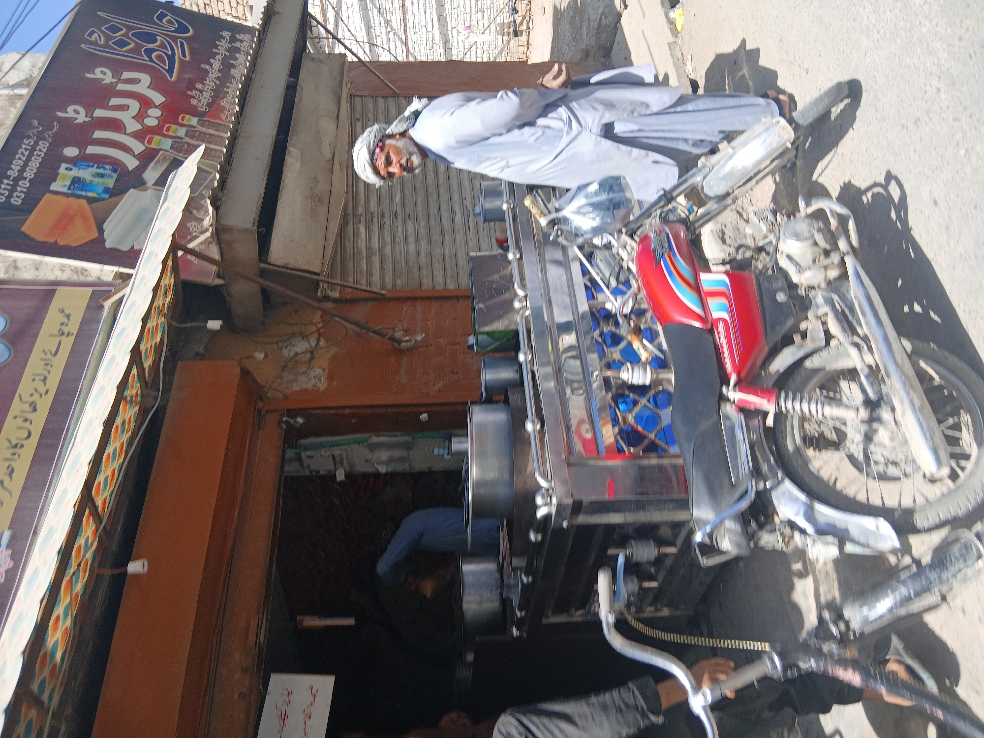 A man in traditional Pakistani clothing stands next to a decorated motorcycle rickshaw in Quetta City Tehsil, Pakistan. The man wears a white shalwar kameez and a turban. The rickshaw is brightly colored and features intricate metalwork. The scene is captured in front of a building with shops and signage in Urdu. The environment appears to be a bustling urban street.