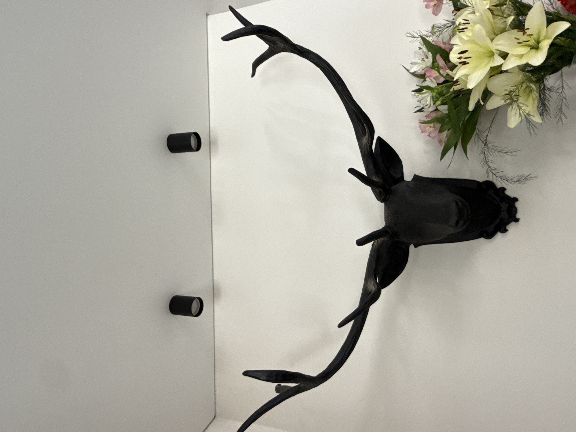 A black, stylized deer head mount with prominent antlers is displayed on a white wall. Two black cylindrical lights are mounted on the ceiling above the deer head. To the right of the deer head, a bouquet of flowers, primarily white lilies with hints of pink, is arranged in a vase. The setting appears to be an indoor space, possibly a home or a public building, in the city of Khokhryaki, Russia.