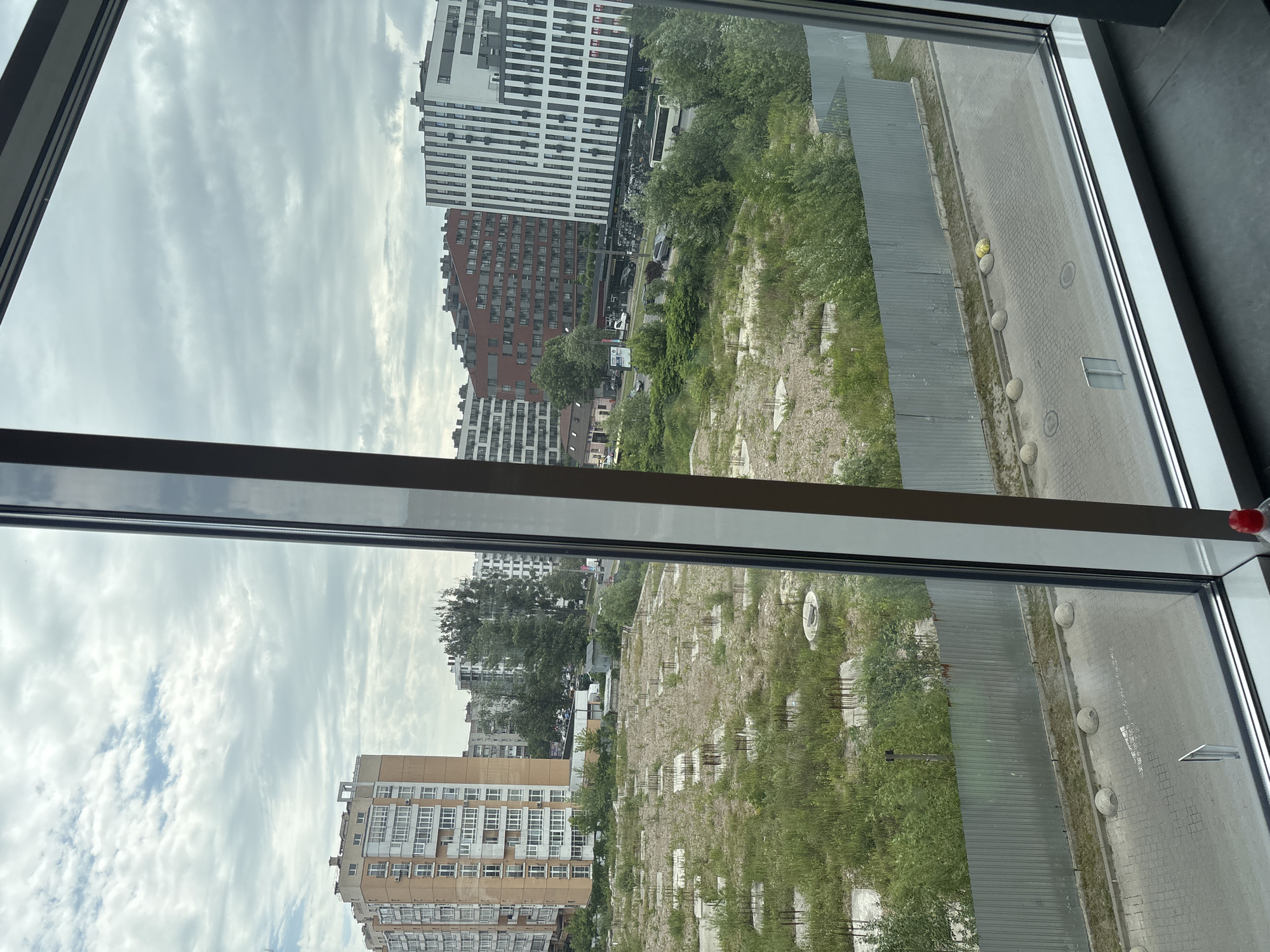 A medium shot captures a view through a window overlooking a city scene under a cloudy sky. In the foreground, the window frame is visible on the right and left sides of the image, with the bottom sill of the window extending across the lower portion. Outside the window, a construction site is present with numerous concrete pillars and rebar protruding from the ground, covered in overgrown grass and weeds. A corrugated metal fence encloses the site. Beyond the construction area, a paved road with bollards and a white arrow painted on its surface is visible. Further back, several modern buildings of varying architectural styles and heights are situated, some featuring brick facades and others with white and black striped exteriors. Trees and foliage are interspersed among the buildings. Cars and a bus are observable on the road in the middle ground. The sky above is filled with a mix of white and gray clouds.