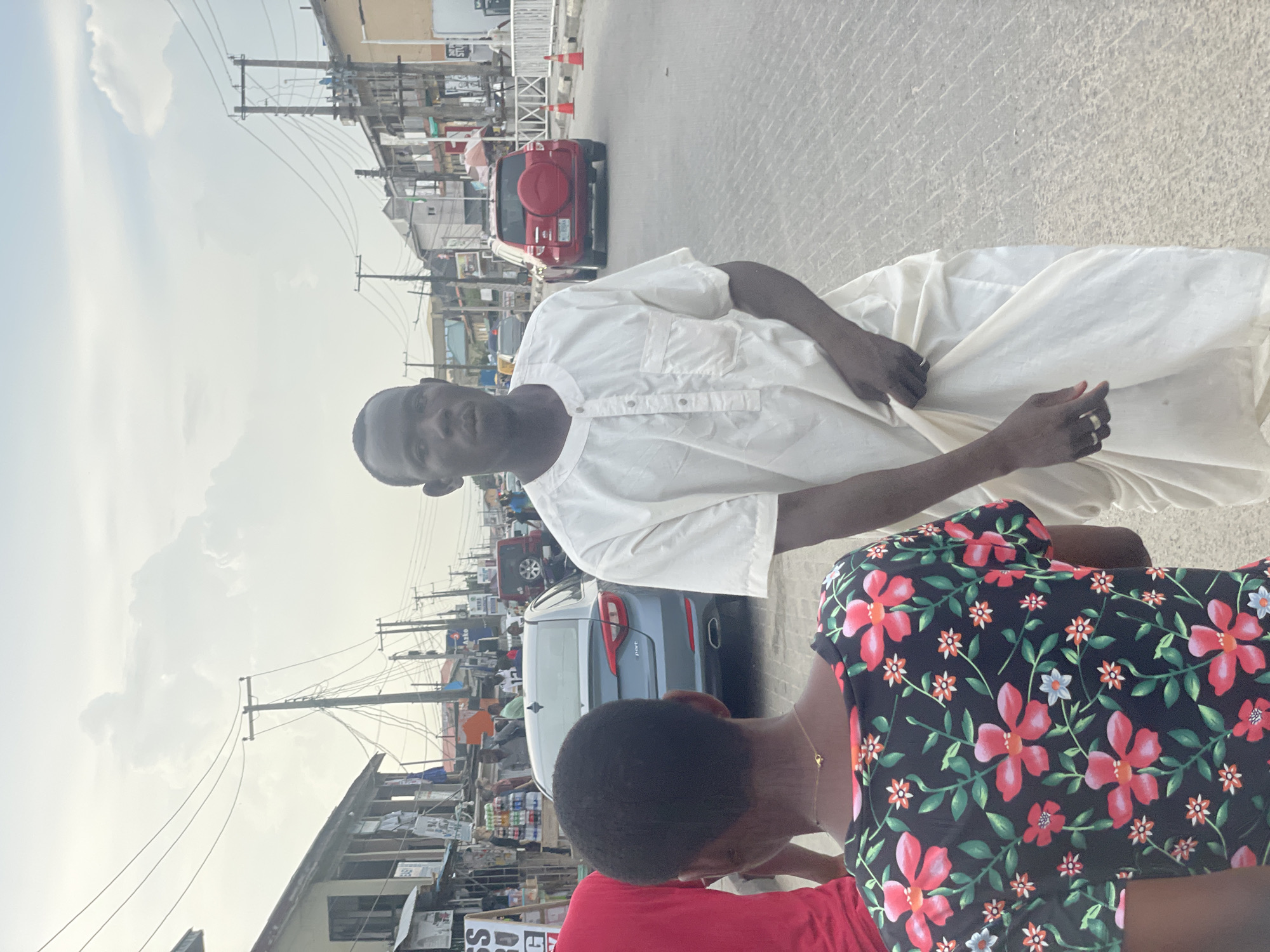This media file captures a vibrant street scene in Ado, Nigeria, under a bright, cloudy sky. In the foreground, a dark-skinned man, dressed in a traditional long-sleeved white tunic (resembling an agbada), stands facing the camera with a neutral expression. He clasps his hands in front of him, and rings are visible on the ring fingers of both hands. Partially visible to his right, with her back to the camera, is another person wearing a dark top adorned with a prominent red and green floral pattern, layered over a red garment.
The background depicts a bustling urban street. A red SUV with a rear-mounted spare tire is parked or moving in the mid-ground, followed by a blue sedan further down the road. The street is lined with numerous utility poles supporting a dense network of power lines. Adjacent to the road are buildings that appear to be shops or market stalls, some displaying indistinct signs. One sign in the distance appears to read "AWO". The road surface is paved with patterned interlocking stones. The bright light suggests it is daytime, likely late morning or early afternoon. Traffic cones are also visible on the roadside further away. The overall scene reflects the dynamic atmosphere of a commercial or residential street in a Nigerian city.