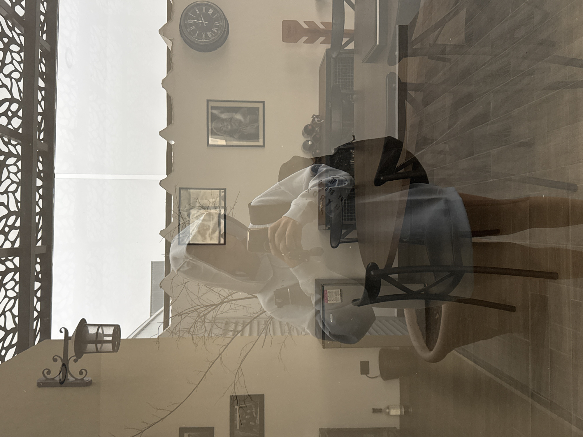 The image is a double exposure of an interior and an exterior scene. The interior features a clock, artwork, and decorative objects on a wall. A table and chairs are visible, suggesting a dining area. The exterior scene shows architectural details of buildings, bare tree branches, and a wall sconce. The overall impression is a superimposed, somewhat surreal juxtaposition of interior and exterior elements. The location mentioned (San Bernardino Tlaxcalancingo, Mexico) does not appear directly deducible from the image content.