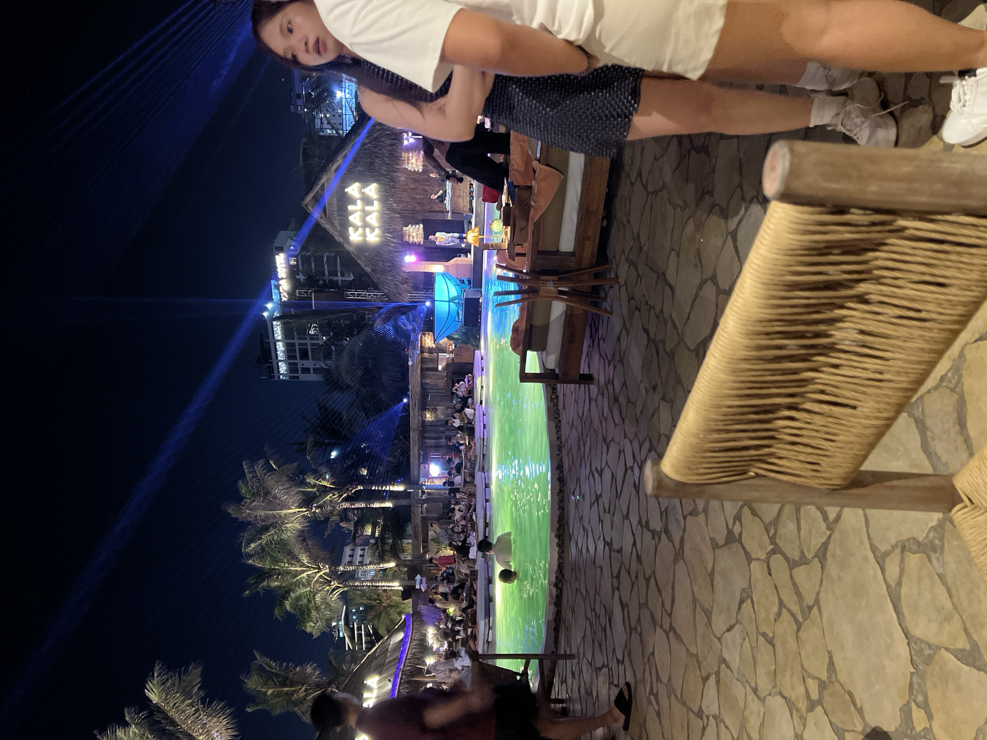 This is a nighttime image of the Kala Kala beach club and entertainment venue located in Đà Nẵng, Vietnam. In the foreground, a portion of a woven chair is visible. The mid-ground features a brightly lit, neon-green swimming pool where two individuals are present. Surrounding the pool are lounge areas with seating, and further back, a thatched-roof structure displays the "KALA KALA" logo illuminated by yellow lights. Palm trees are scattered throughout the scene, and in the background, several multi-story buildings and the night sky with visible blue light beams from a laser show are present. Several people are observed in the background, some seated at tables and others standing. Two women are in the right foreground, partially obscuring the view of the pool area.