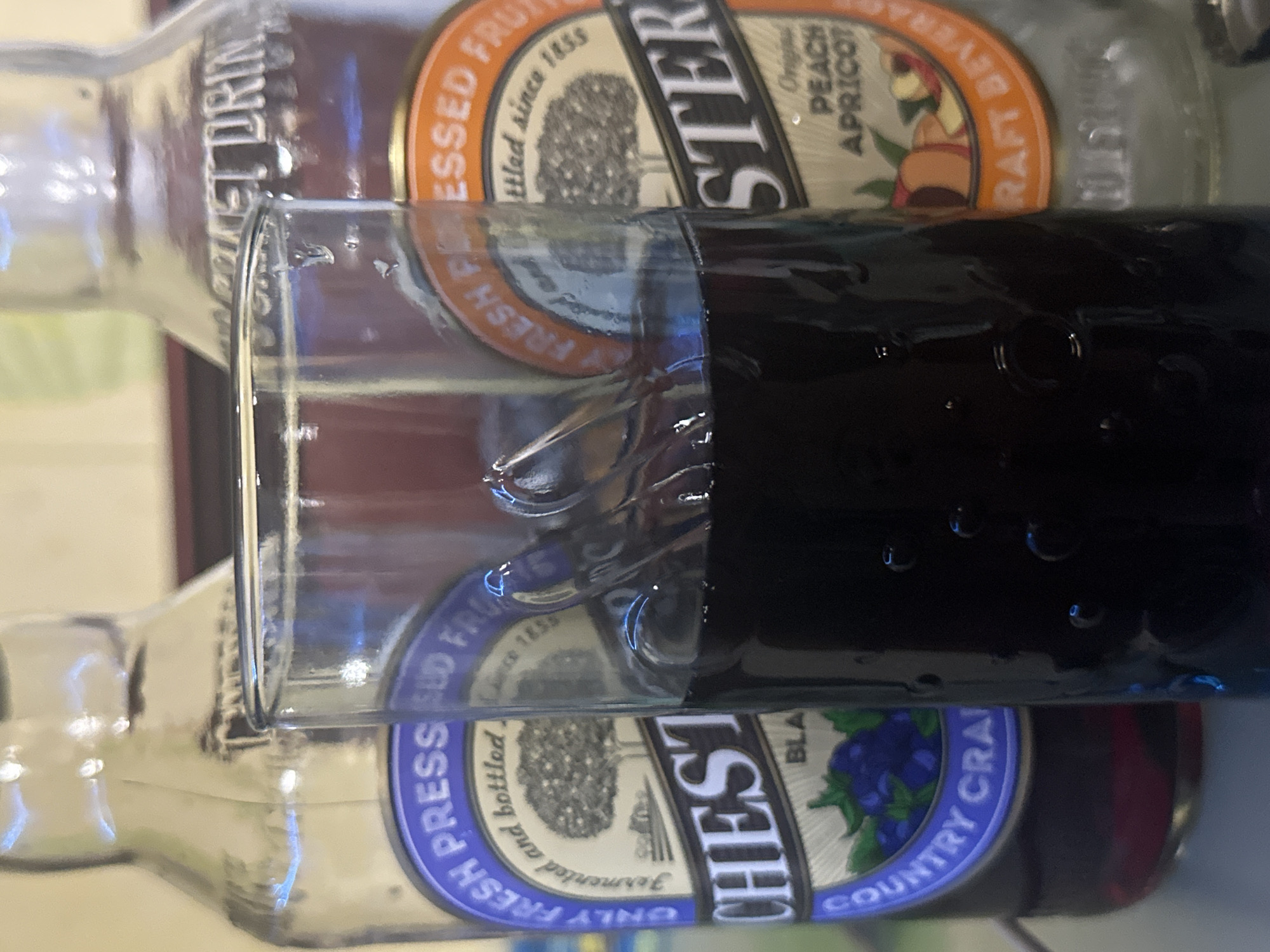 The image shows two bottles of "CHESTER" brand fruit drinks, one labeled "PEACH APRICOT" and the other "BLACK COUNTRY CRAI." A glass filled with a dark liquid is positioned in front of the bottles, partially obscuring their labels. The scene appears to be indoors, but there's no information to pinpoint the location in Moscow, Russia.