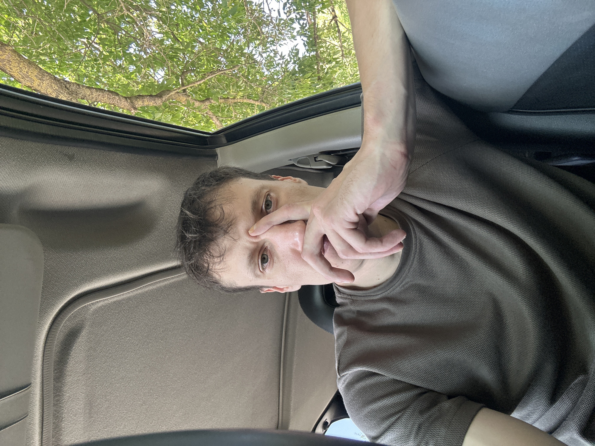 A man is in the driver's seat of a car. He is wearing a brown t-shirt and has his right hand over his mouth and nose. His left arm is resting on the car door. Sunlight is shining through the trees outside the car. The scene is in Makiv, Ukraine.