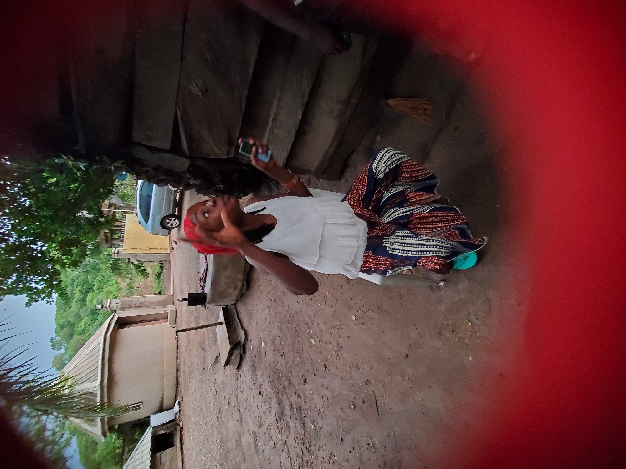 A person, likely female, in a white top and patterned skirt is seated on a dirt ground outdoors, holding a blue smartphone. She is looking up at the phone. A second person's feet are visible behind her. The setting appears to be a rural area with buildings, trees, and a parked car in the background. The photo has a red, blurry border. The location is suggested to be Mararaba, Nigeria, based on the prompt.