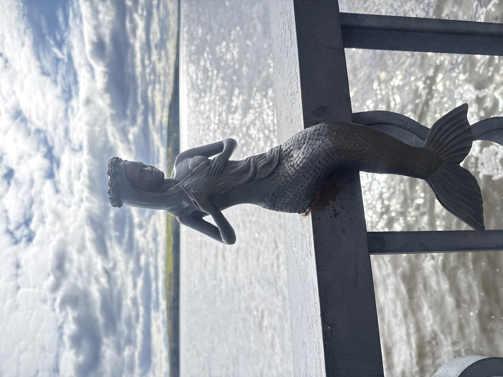 A statue of a mermaid is affixed to a metal structure. Behind the statue, there is a body of water and a cloudy sky. The statue is positioned on a bridge-like structure in the city of Chaikovsky, Russia. The statue is gray and depicts a reclining mermaid.