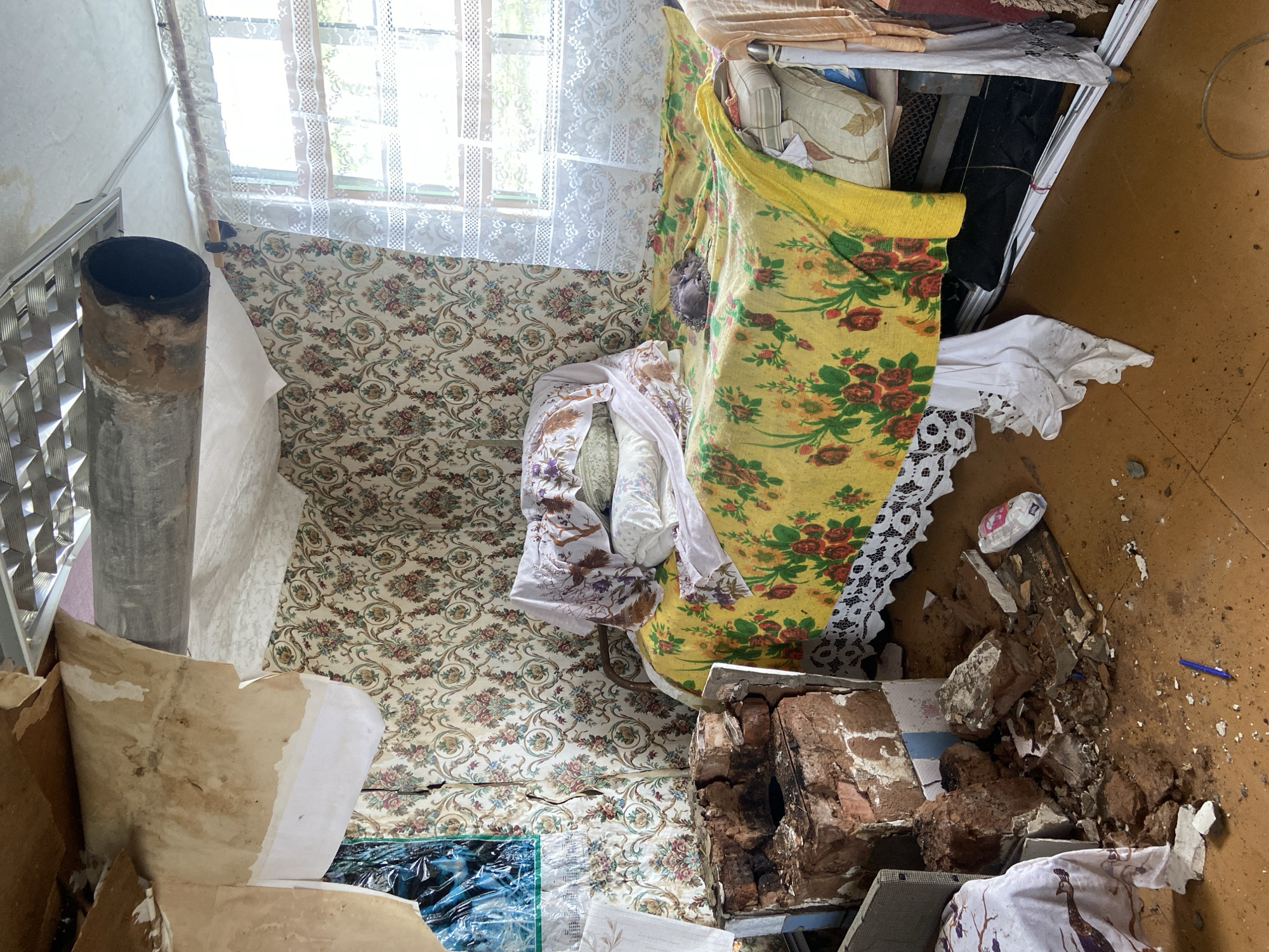 The image depicts a room in disarray, likely located in Dubovaya, Russia. The walls are covered in floral wallpaper, partially peeling. A window with lace curtains is visible. A rusty stovepipe stands near the wall. Debris, including broken bricks and fragments of materials, litters the floor. A bed, covered with floral patterned sheets and pillows, is partially draped with a yellow floral fabric. A shelf unit, possibly made of metal, stands against a wall, containing various items. The floor is a wooden surface. The overall impression is one of abandonment and decay.