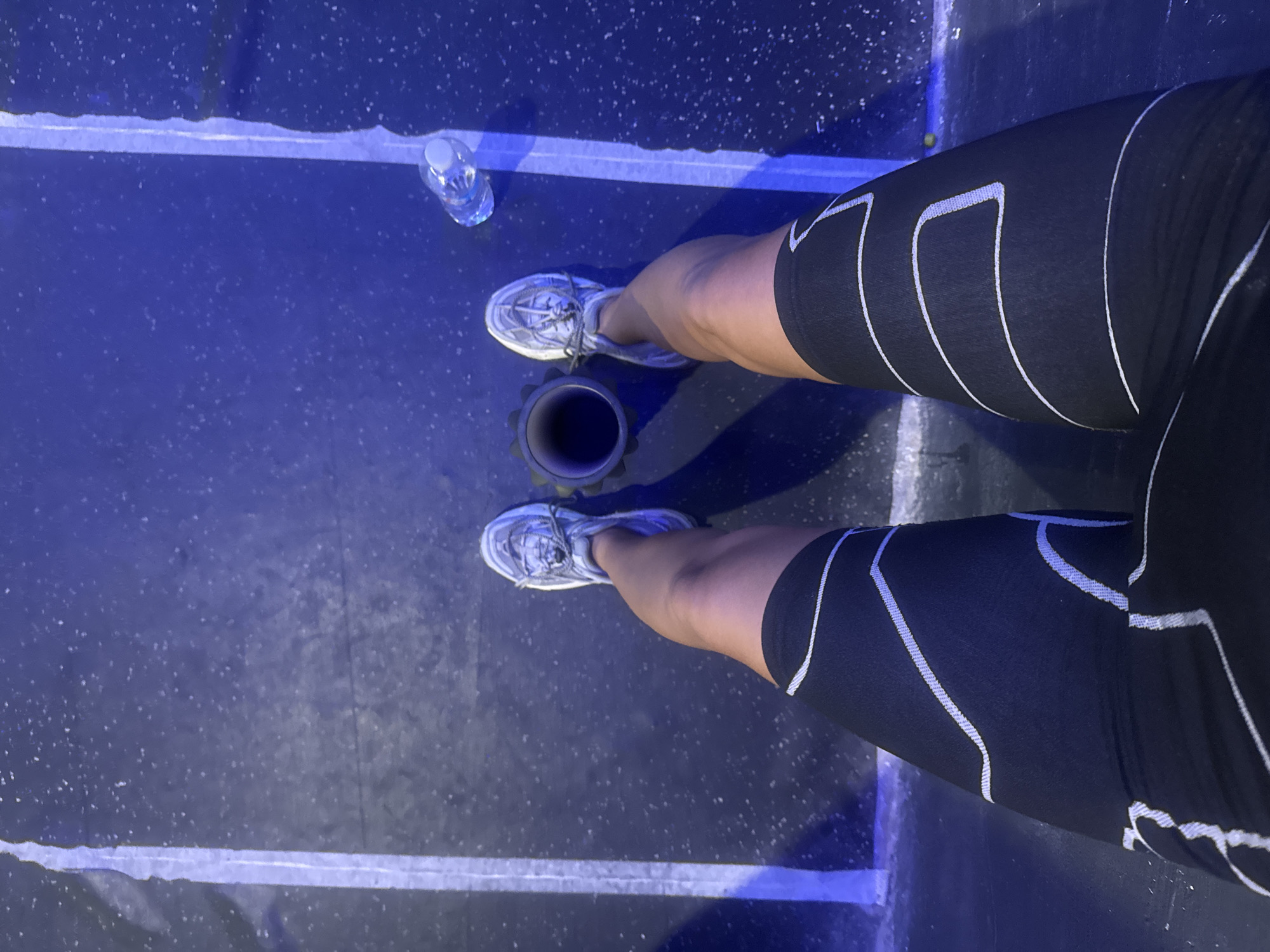 A downward-angle, medium shot shows a person's legs, wearing athletic shoes and compression shorts, on a dark, possibly rubberized floor with white lines suggesting a fitness or sports area. A water bottle and a black cylindrical object (possibly a foam roller) are also present on the floor. The lighting is somewhat dim with a bluish tint. The location in the city of Đà Nẵng, Vietnam is indeterminable from the image.