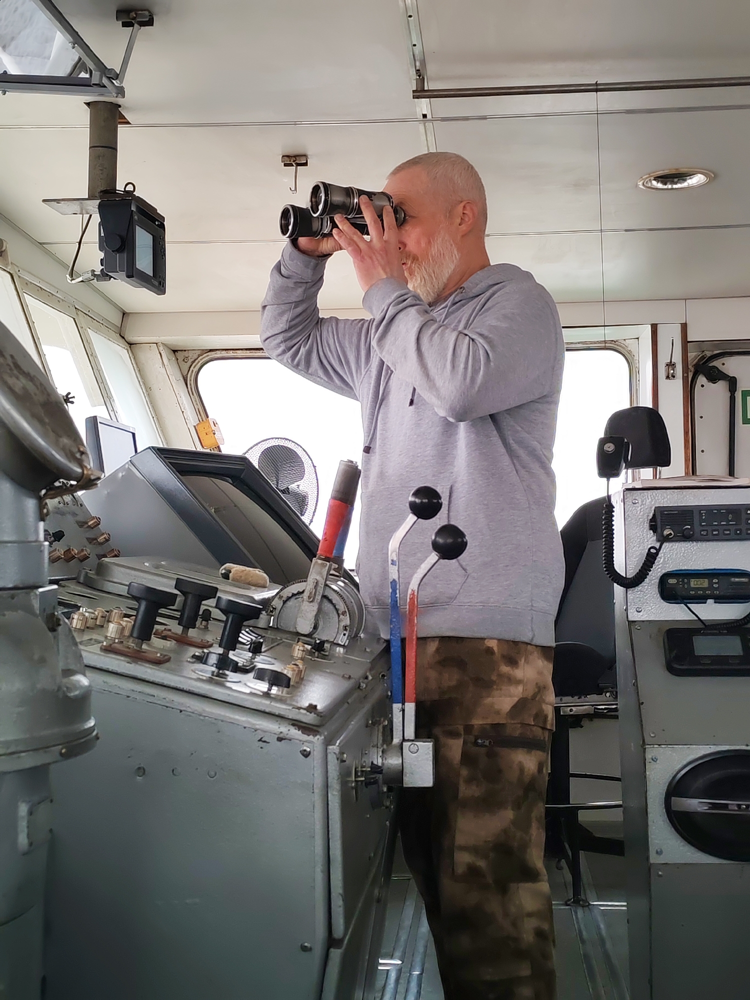 Inside the functional bridge of a ship, a man with a graying beard and short hair, wearing a gray hooded sweatshirt and camouflage pants, stands facing right, looking intently through a pair of black binoculars. His hands are raised to hold the binoculars to his eyes, suggesting he is actively scanning the surroundings or horizon.
The ship's interior is utilitarian, with light-colored walls and ceiling, and robust gray control panels. To the man's left is a large main console featuring numerous dials, buttons, and levers, including two prominent control handles with red, white, and blue markings, indicating engine or steering controls. Above this console, a square display unit, possibly for navigation or radar, is mounted from the ceiling on a metal pole.
Bright, diffused light streams in through multiple large windows along the front and side of the bridge, suggesting daytime, possibly under an overcast sky or in foggy conditions. To the man's right, a separate console holds communication equipment, including a radio with a coiled microphone and a digital display showing "002". A small, circular fan is visible behind the main console. A green symbol, likely a safety or exit indicator, is partially visible on the right wall. The overall scene conveys a sense of operational readiness on a vessel, likely in the area of Shcheleiki, Russia.