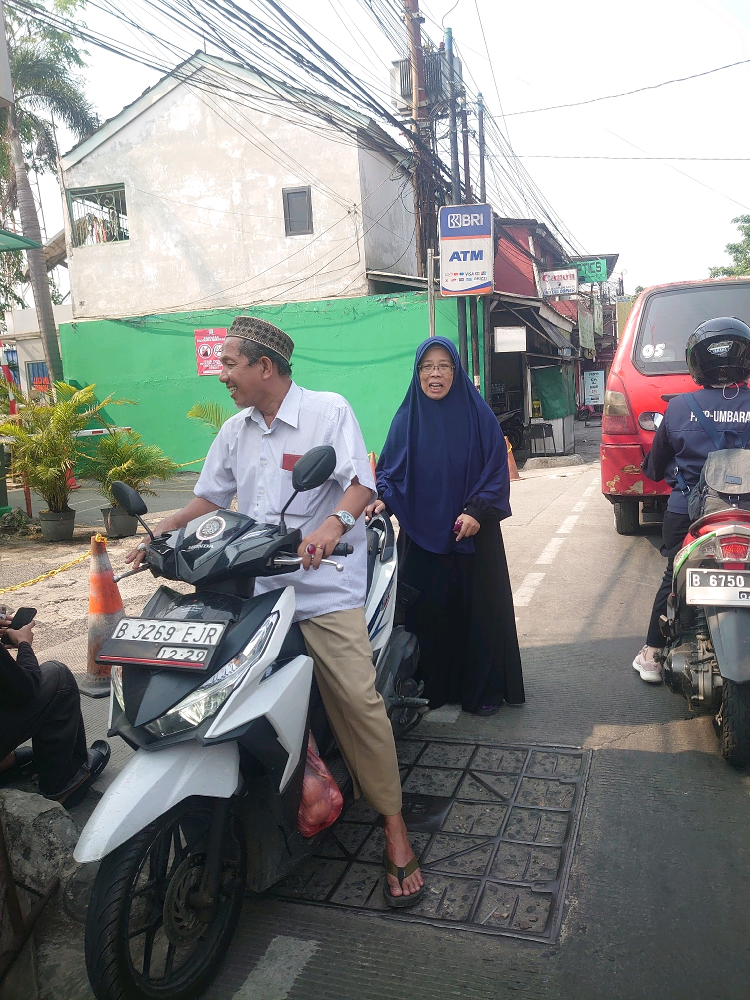 In Bojong Gede, Indonesia, a man wearing a patterned cap and a light-colored short-sleeved shirt is seated on a white Honda motorcycle, looking to his left and smiling. Beside him, a woman in a navy blue hijab and a long black dress stands facing the viewer with her mouth open, seemingly speaking or reacting. The motorcycle has a license plate that reads "B 3269 EJR" and "12-29". To the right of the couple, a person in a black helmet and a blue jacket is on a motorcycle. In the background, a red truck is parked. The environment includes a green wall, a building with a white facade and windows, a transformer with numerous wires, and signs including a BRI ATM sign and a "Canon" sign. The ground is paved, and a metal grate is visible in the foreground. Traffic cones are also present.