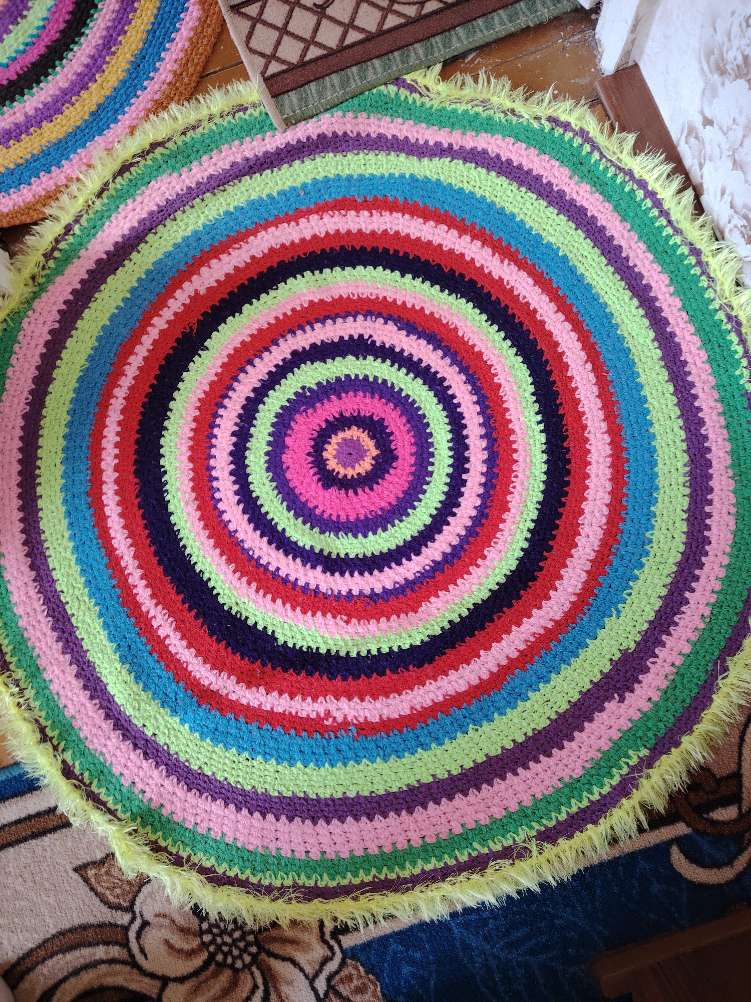 This image presents a vibrant, circular, handmade crocheted rug, prominently displayed on a floor in what appears to be an indoor setting. The rug features a striking pattern of concentric rings, crafted in a rich palette of bright colors including various shades of green, blue, pink, purple, and red. A distinctive detail is the fluffy, bright yellow fringe that outlines the entire circumference of the rug.
In the top-left corner, a fragment of a second, similar multicolored crocheted rug is visible, suggesting a coordinated set or multiple handmade pieces. The floor beneath and around the main rug is covered by other patterned carpets or rugs; one section visible in the background is a brown and beige carpet with a traditional floral or geometric design, while another area shows a blue carpet with darker patterns. Parts of a wooden floor or surface are also discernable.
No individuals are present in the image, and no specific activity is occurring. The photograph captures a static arrangement of home decor, illuminated by what appears to be bright, ambient daylight. The overall impression is one of handcrafted warmth and cheerful aesthetics. There is no visible text within the image. The location context for this scene is Yanaul, Russia.