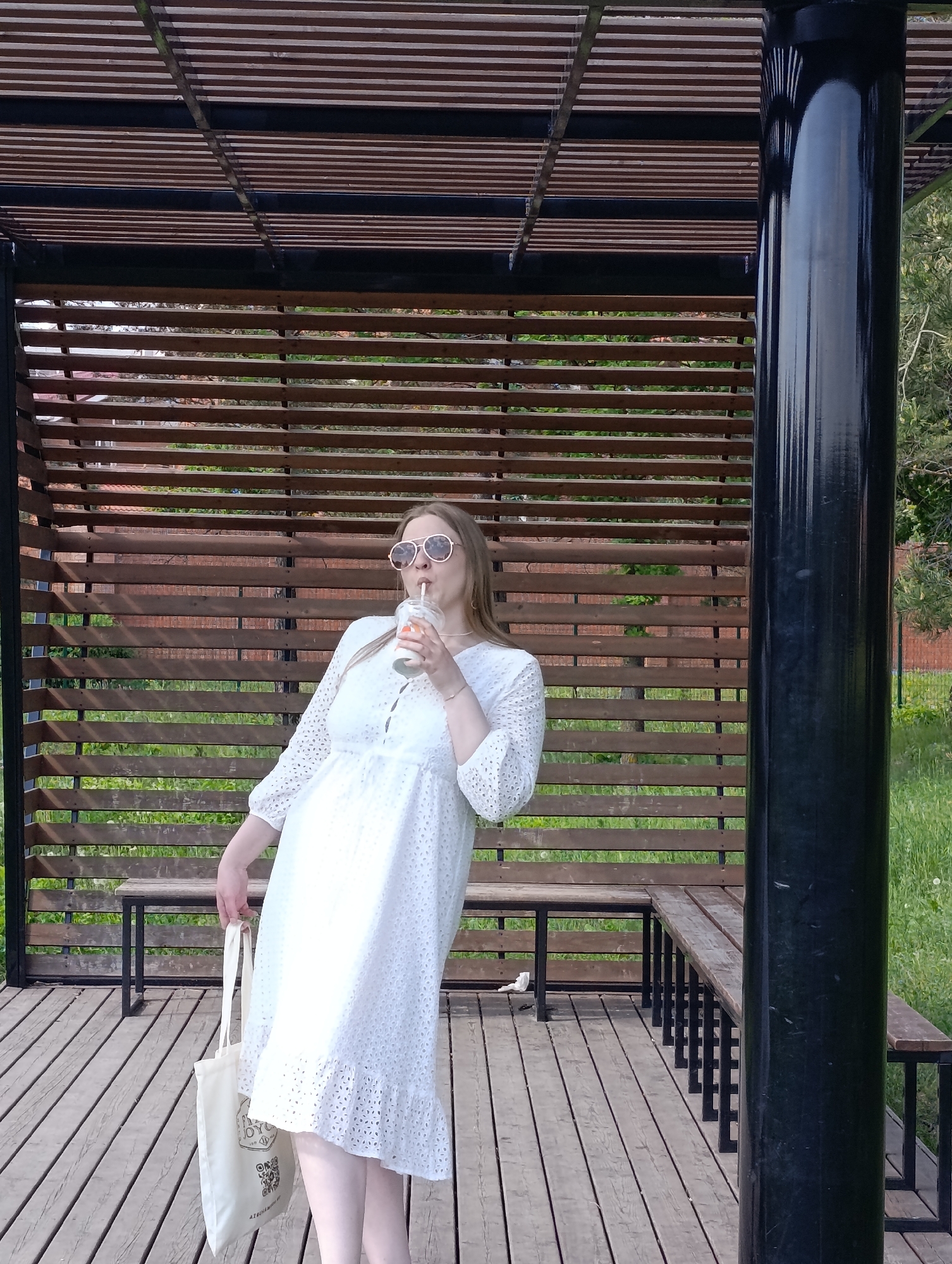 A full shot captures a woman in a white dress standing in a gazebo, drinking from a cup with a straw. She wears round sunglasses and holds a tote bag. The gazebo has a wooden slat backdrop, a slatted roof, and benches with black metal frames. The flooring is wooden planks. A black pole is on the right side of the frame. Outside the gazebo, there's green grass and glimpses of trees.