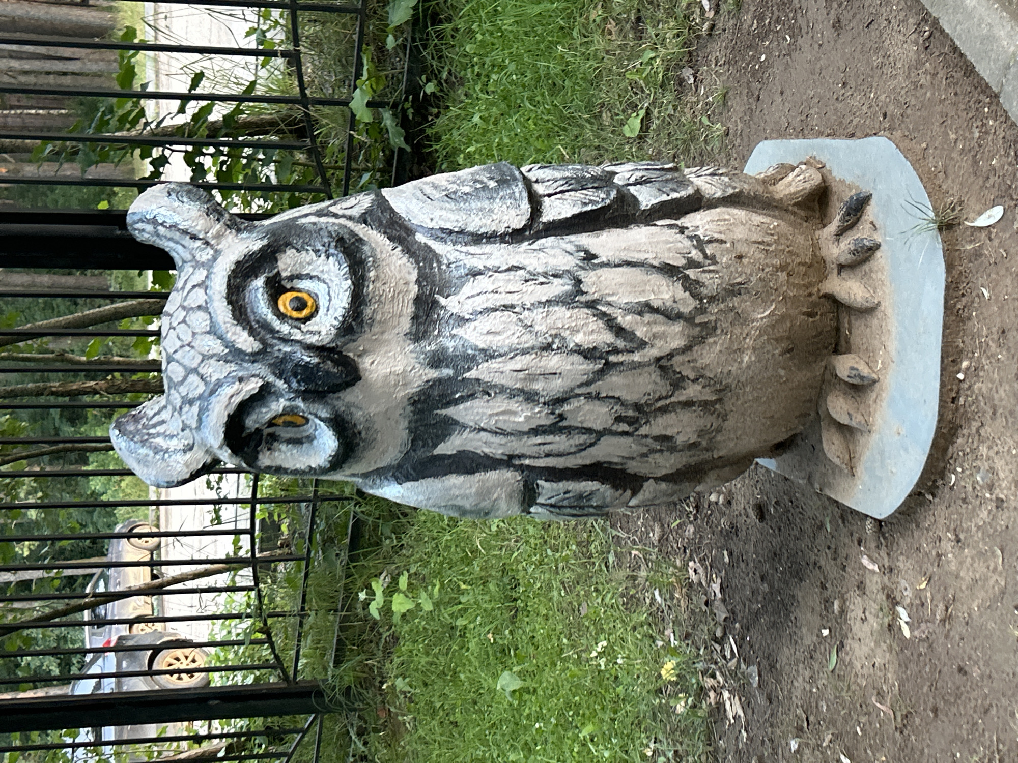 A statue of an owl, painted gray and black with yellow eyes, stands on a concrete base on a dirt and grass patch. Behind the statue, a black metal fence separates the area from a paved surface where two dark-colored cars are parked. Lush green grass and trees are visible beyond the fence. The scene appears to be an outdoor location in the city of Chaikovsky, Russia.