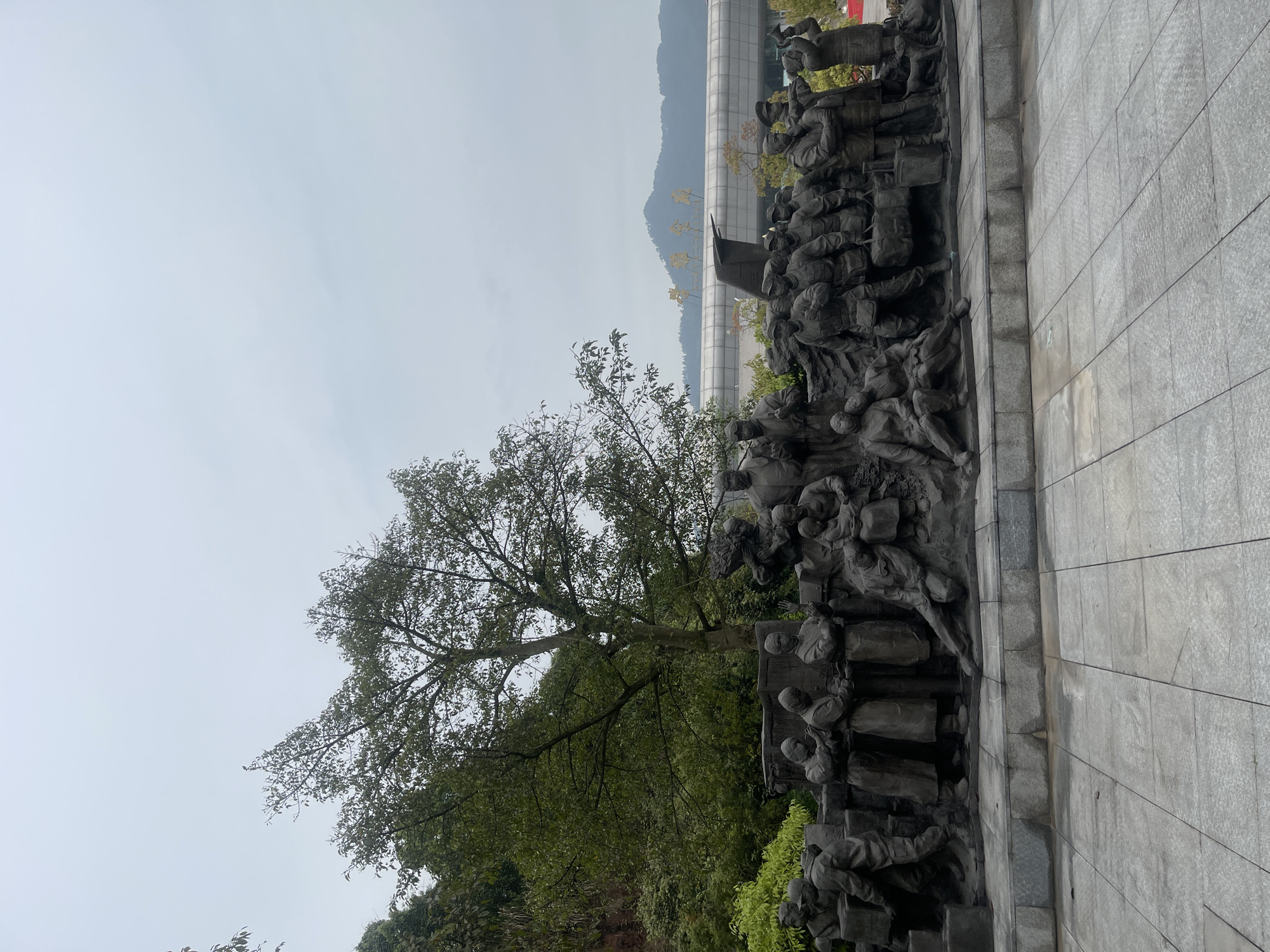 The image shows a section of a bronze relief sculpture located in the Chongqing High-tech Zone, China. The sculpture depicts a scene of marching figures with a flag, possibly soldiers or revolutionaries. A large tree is situated next to the sculpture, and a building is visible in the background, partially obscured by distant mountains and a cloudy sky. A gray stone surface makes up the foreground.