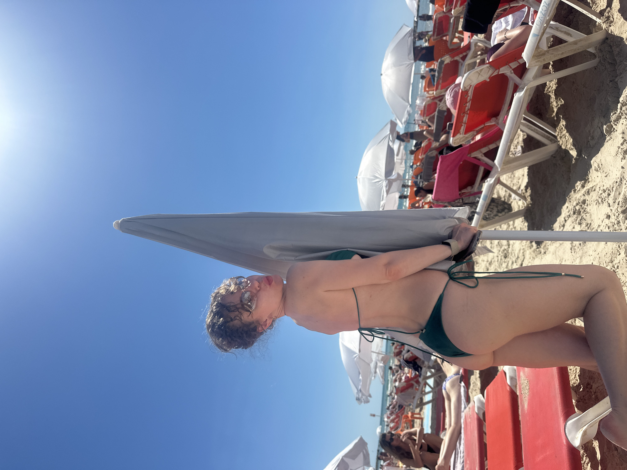 The image depicts a woman in a green bikini lying on a red beach chair under a white umbrella on a sunny day. The scene is at a beach with numerous other beach chairs and umbrellas, likely crowded with people. The sky is a clear blue. The location is likely a beach in Tel Aviv, Israel.