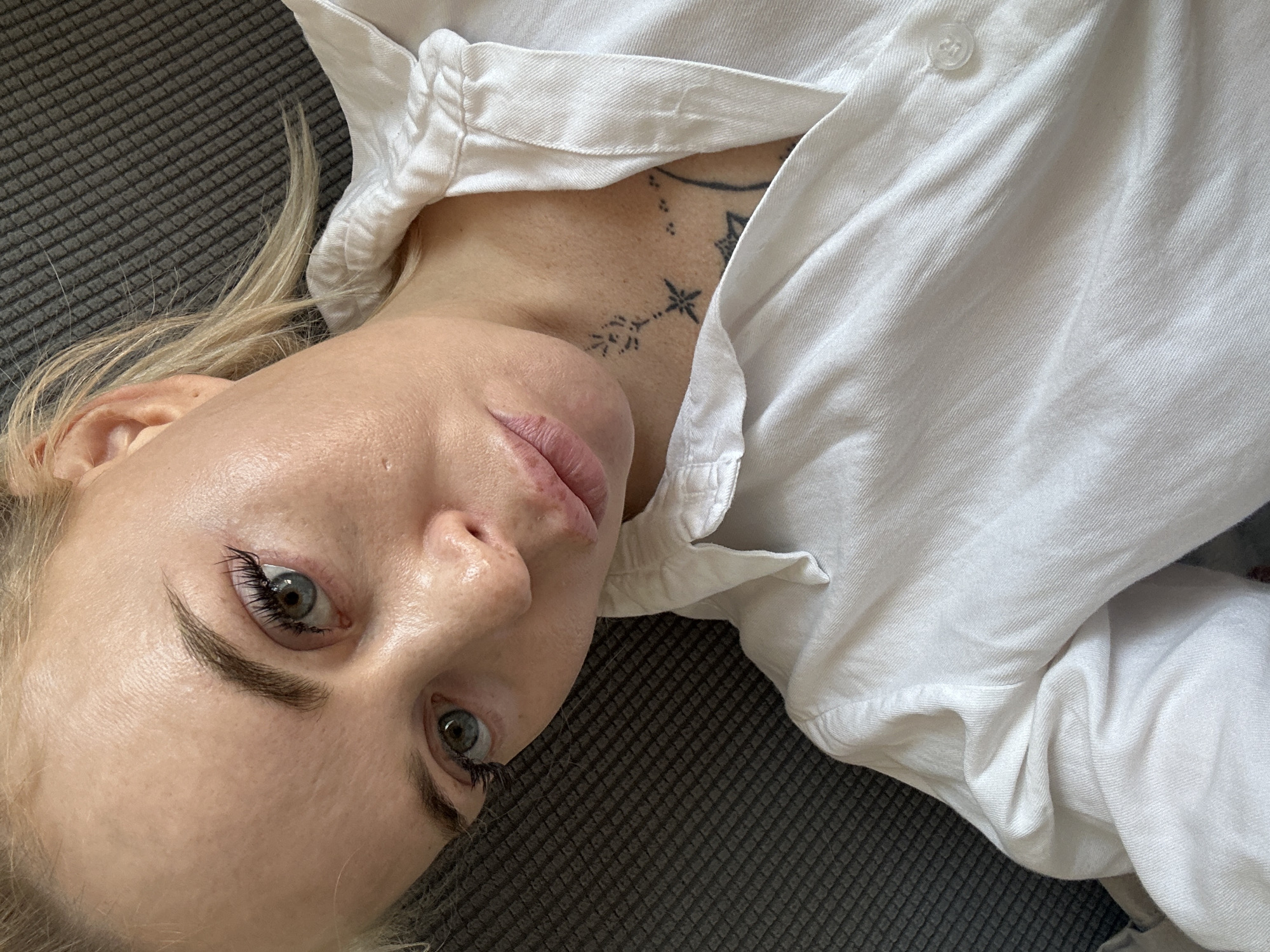 An overhead, close-up, indoor shot shows a fair-skinned woman with blonde hair pulled back in a ponytail, wearing a white button-down shirt and lying on a dark gray, textured surface. The woman's face is turned to the left, with her eyes open and looking towards the camera. She has light blue eyes and wears mascara. Her lips are slightly parted. A small, dark tattoo is visible on her neck, just above her collarbone. The shirt has a folded collar and buttons down the front. The background is out of focus.