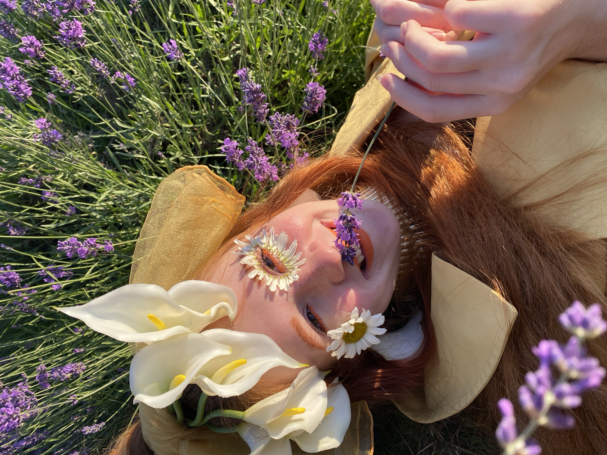The media file shows a close-up, overhead view of a person lying in a field of lavender, adorned with flowers. The person has red hair and is wearing a golden, wide-brimmed hat. White calla lilies are placed around their face, and daisies are positioned over their eyes and nose. Sprigs of lavender are also woven into the scene, with some placed near the person's face and others in the surrounding field. The person's hands, with manicured nails, are visible in the upper right corner, resting near their face. The setting is outdoors, with lush green foliage and vibrant purple lavender blossoms. The lighting suggests it is daytime, likely sunny, given the highlights on the hair and skin, and the overall brightness of the scene. The overall mood conveyed is serene and ethereal, with a strong connection to nature. The location context provided is Verkhneye Uchdere, Russia, which aligns with the presence of lavender fields. There is no visible text in the image.