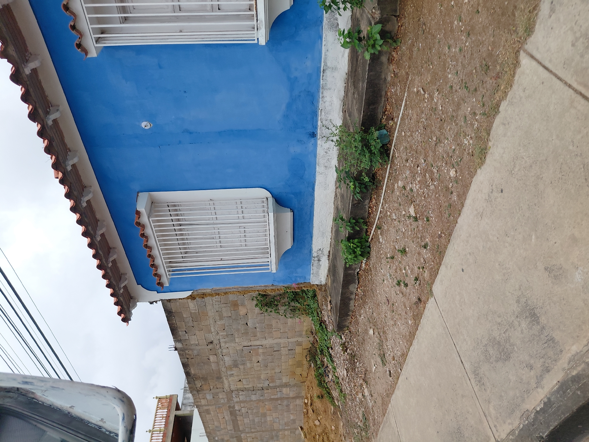 The image shows a two-story building in Altagracia, Venezuela. The top story is painted bright blue with white trim around the windows and roofline, which has red roofing tiles. The bottom story appears to be constructed of cinder blocks. The sidewalk is visible on the right of the image, and the ground between the building and the sidewalk is bare dirt with sparse vegetation. A vehicle is partially visible in the bottom left corner. Power lines are visible above the building.