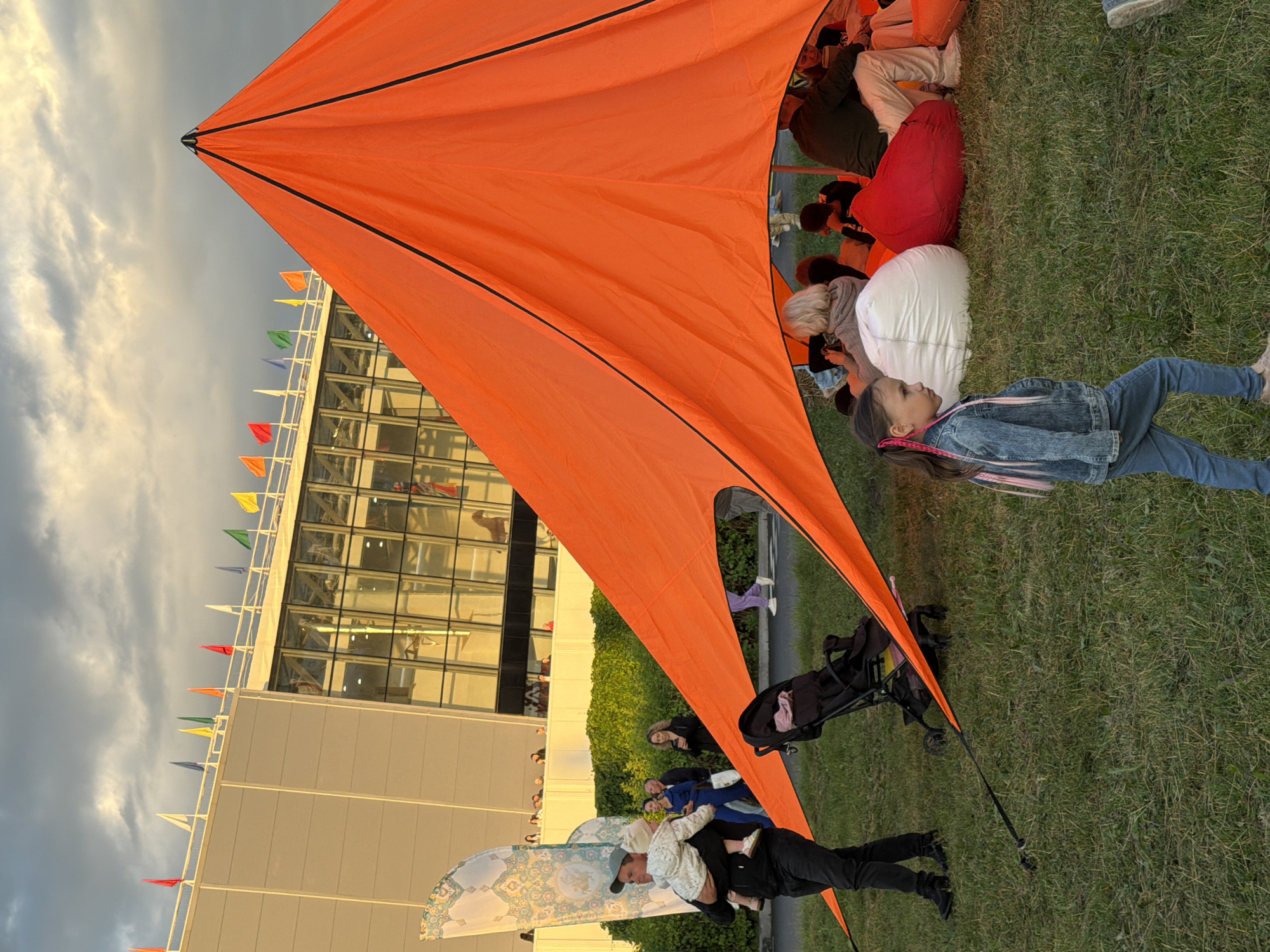 An outdoor scene depicts a gathering under a large, bright orange tent. The tent is a prominent structure, its fabric stretched over poles and secured to the ground with ropes. Inside the tent, several people are seated on large, colorful beanbag chairs, appearing relaxed. In the foreground, a young girl with braided hair walks across a grassy area. To the left of the tent, a man is holding a baby, and a stroller is visible. In the background, a modern building with a glass facade stands tall, adorned with a row of colorful flags along its rooftop. The sky above is cloudy, suggesting it might be late afternoon or early evening. The overall environment appears to be a public space or park.
