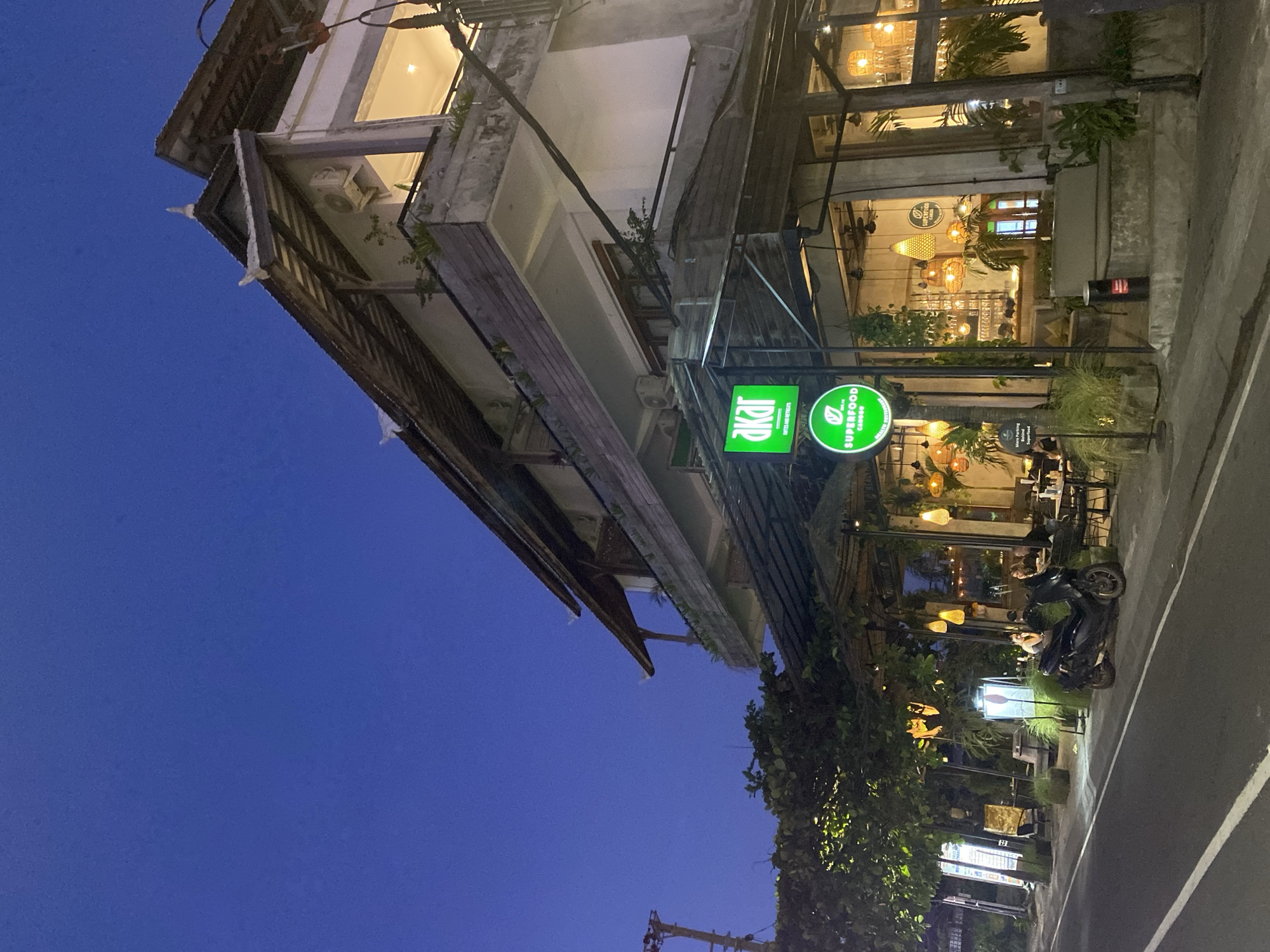 The image depicts the exterior of a building in North Kuta, Indonesia, at dusk or night. The building has a multi-story structure with a mixed architectural style. The exterior features wooden elements, and a lighter colored facade. Illuminated signs for "akar" and "Superfood" are visible near the entrance, indicating a cafe or restaurant. The interior of the establishment has warm lighting and plants. A motorcycle is parked on the adjacent road. Street lights illuminate the area, with trees lining the road. The sky is a deep blue.