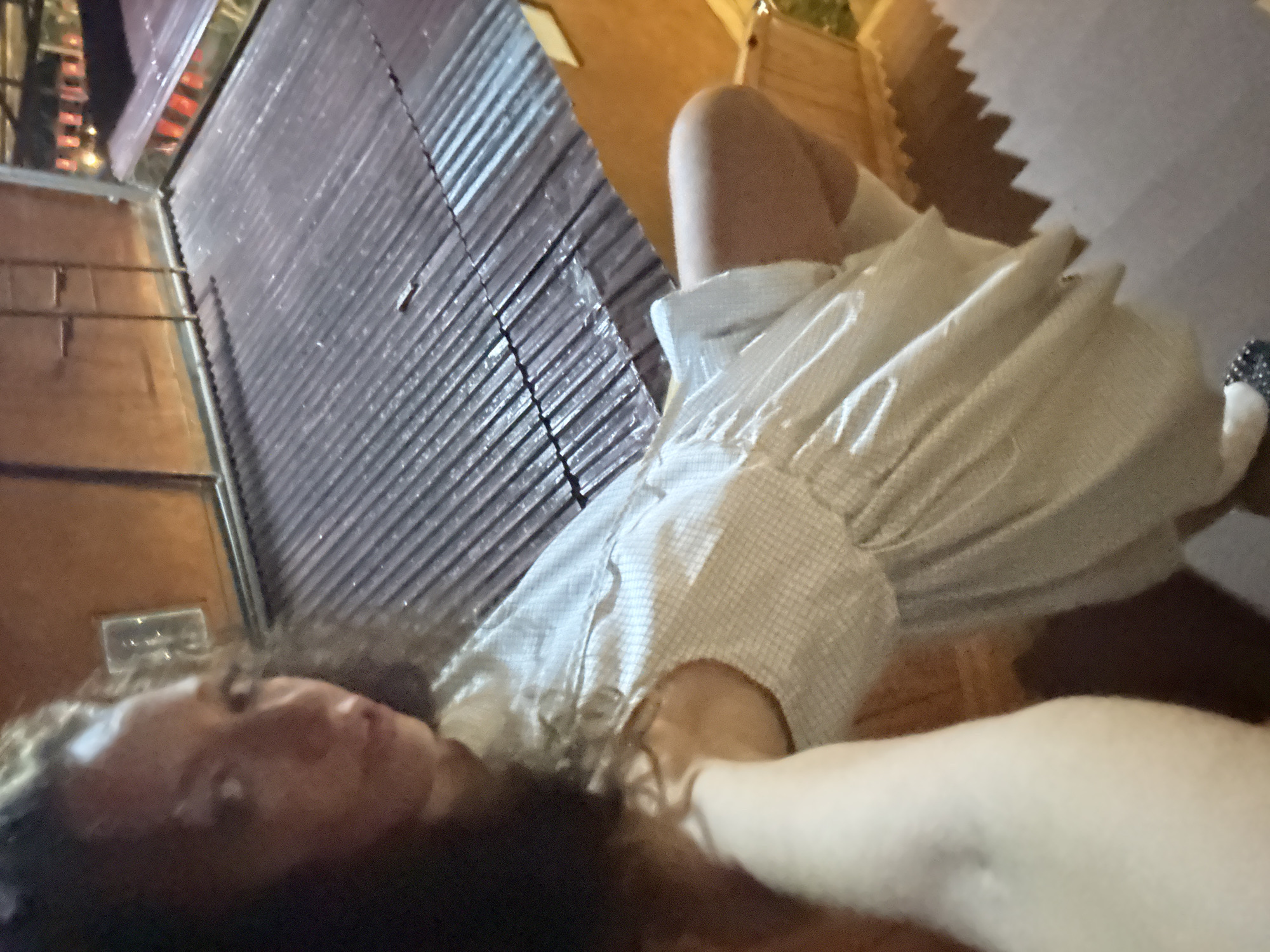 A woman in a light-colored dress is lying down outside, possibly in Bắc Hà, Vietnam. Her head is close to the camera, and her arm is extended towards the viewer. A corrugated metal wall is behind her, and there are wooden structures visible in the background. The scene appears to be at night.