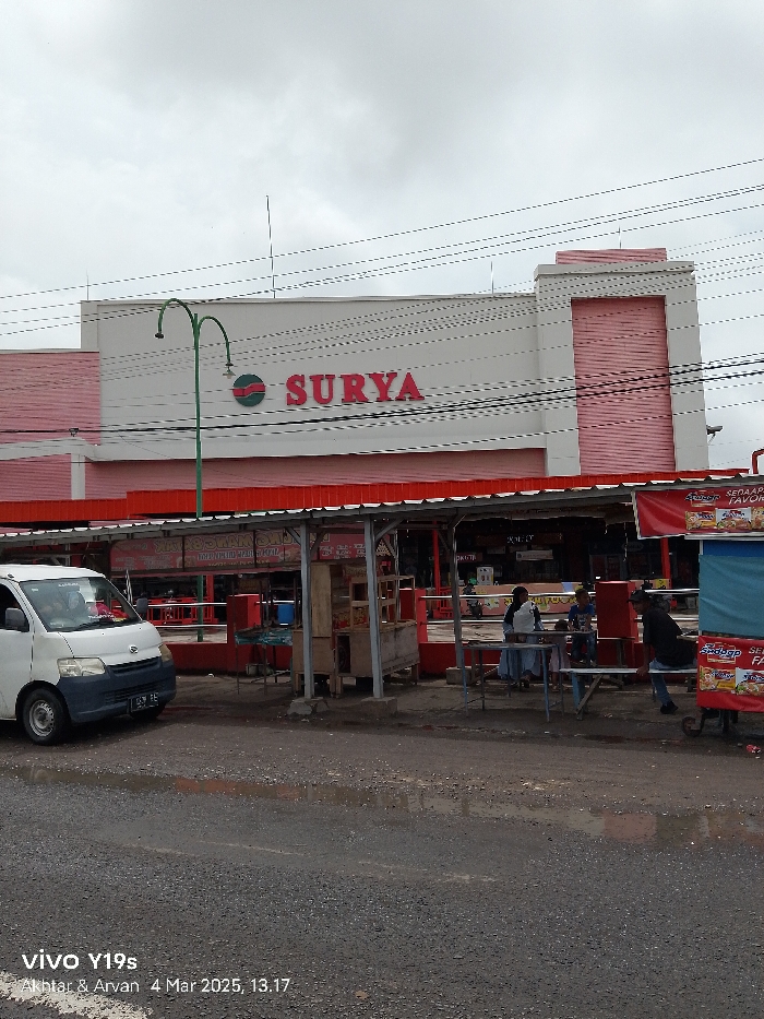 The image shows a street scene in front of a large building with a red and white facade in Jatibarang, Indonesia (latitude: -6.467, longitude: 108.298). The building is a store called "SURYA". A white van is parked in the street in front of the building. In the foreground, there are three people sitting at a table and talking. There is a food stall next to them, and a small cart selling snacks and drinks. There is a red awning overhead. In the background, there is a second food stall with an awning. The street is wet and there are power lines overhead. The image was taken on March 4, 2023, at 1:17 PM.