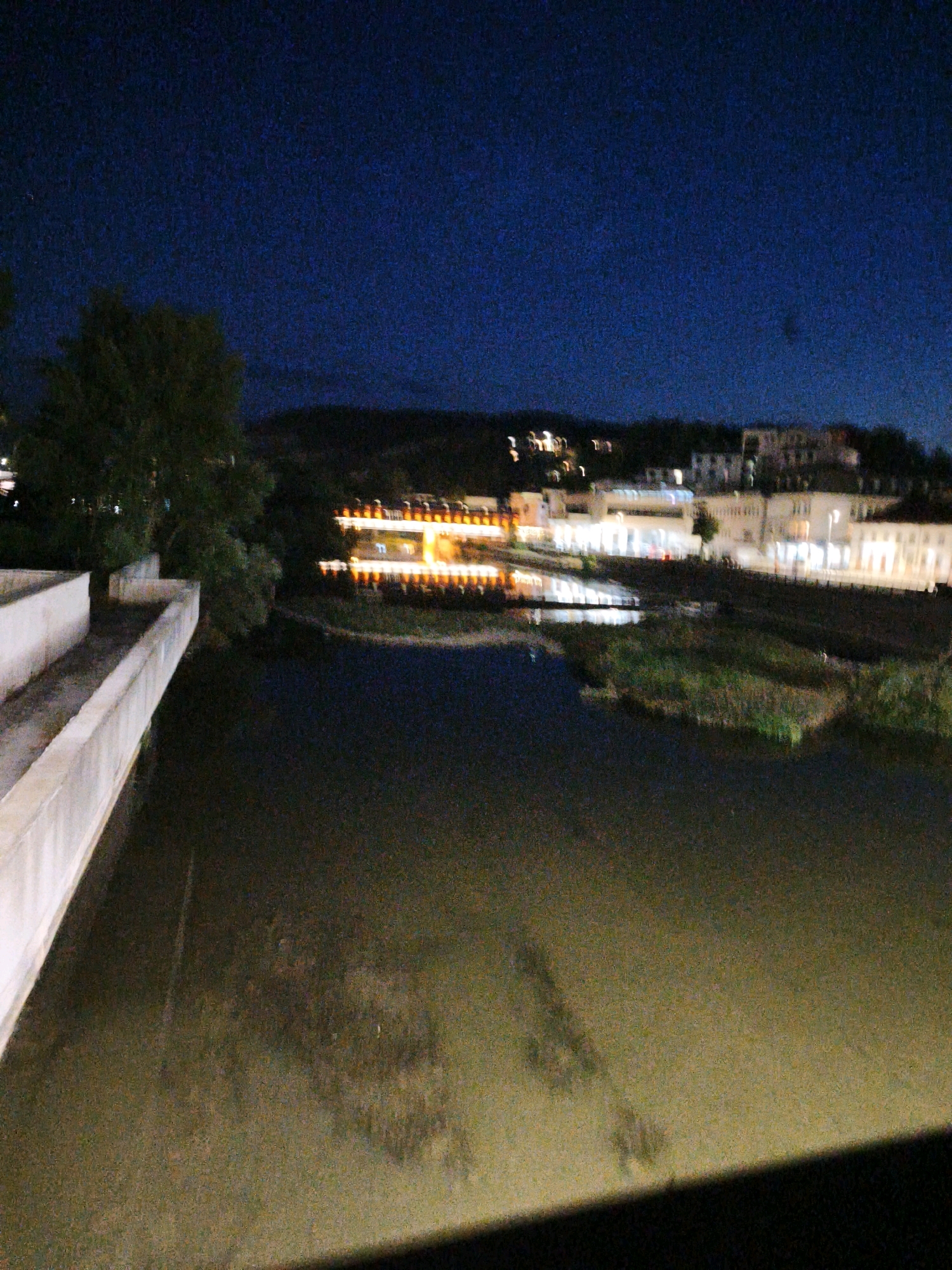 The image depicts a nighttime scene in Lovech, Bulgaria, focusing on the Osam River and its surroundings. In the foreground, a concrete structure, likely a bridge or embankment, runs along the left side of the frame, illuminated by ambient light. The river itself appears dark and reflects some of the distant lights. Across the river, a town or city is visible with illuminated buildings. Distinctive long, horizontal lights are prominent, suggesting a promenade or a building with many windows. A cluster of trees is situated on the left bank of the river, adding a natural element to the urban landscape. The sky is dark, indicating nighttime. The overall impression is of a tranquil evening view of the river and the town.