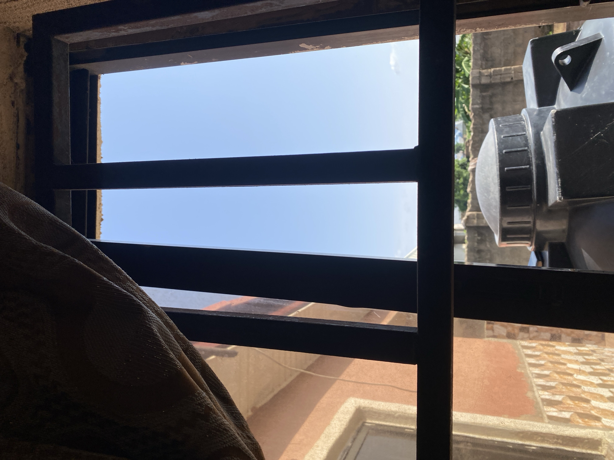 The image shows a view through a barred window on a bright, sunny day. The bars are made of dark metal and are spaced horizontally, allowing a view of the sky and some buildings.
In the foreground, to the left, is a draped piece of textured fabric, possibly a curtain or blanket, in a brown and tan color. This fabric obscures part of the view to the left and bottom of the window.
The sky visible through the bars is a clear, light blue, suggesting fair weather. A few faint clouds are visible.
Through the bars and to the right, a portion of a large black object is visible. It appears to be some kind of tank or container with a textured lid or cap. Further to the right and in the background, parts of buildings are visible. One building has a reddish-orange facade and a tiled roof with a mosaic pattern. Another building in the background appears to be made of concrete. Green foliage from trees can also be seen in the background.
The scene appears to be set in an outdoor environment, likely a residential or urban area, given the presence of buildings and the water tank. The time of day is clearly daytime due to the bright sunlight and clear sky. There are no people visible in the image, and the overall impression is one of stillness and observation. No text is visible in the image. The context provided indicates the location is Jauro Yinu, Nigeria.