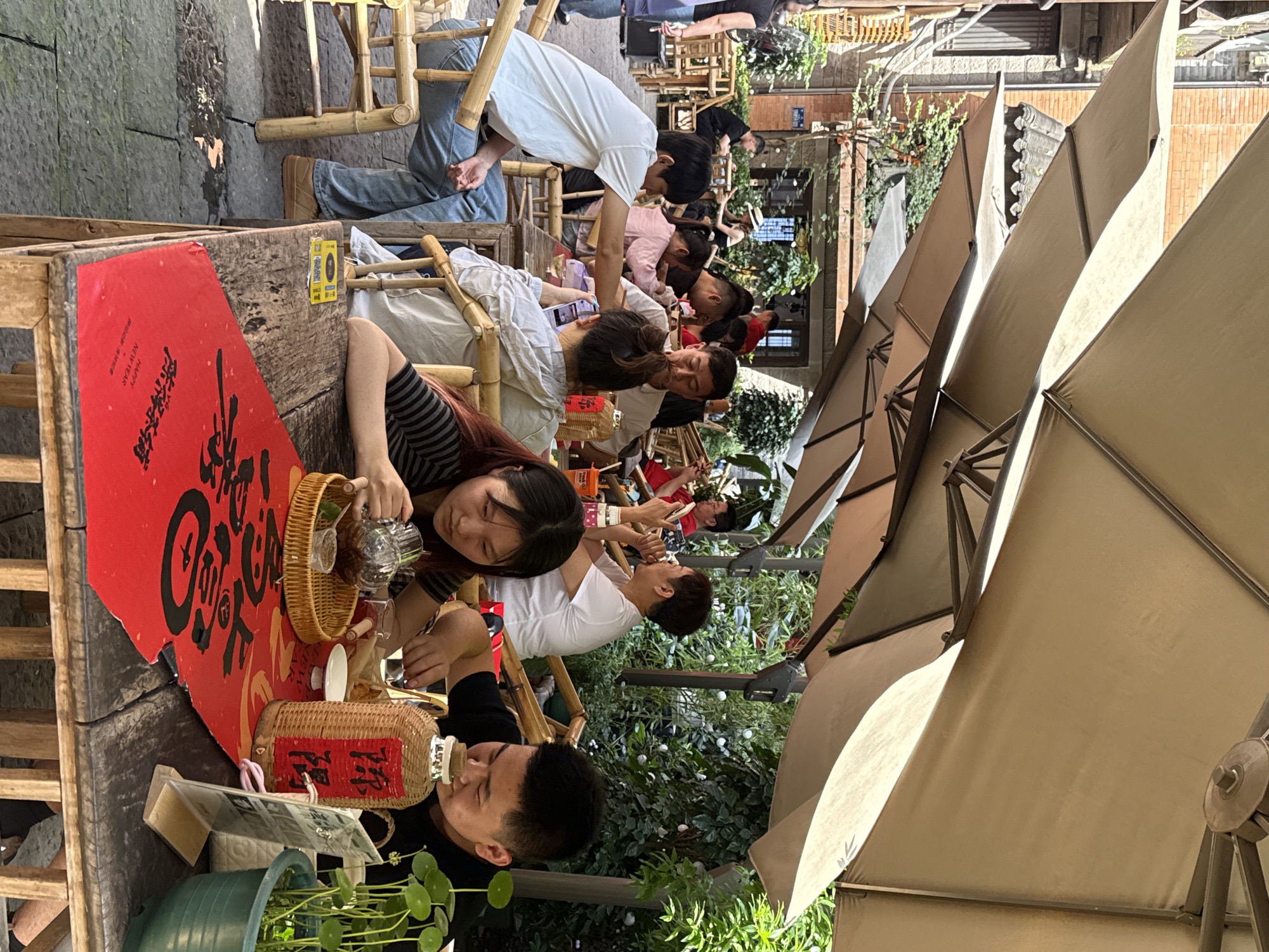 This image depicts a bustling outdoor cafe or tea house scene, likely in Yuzhong District, China, during a bright, sunny daytime. Numerous people are seated at rustic tables and chairs made of bamboo or wood, enjoying beverages and socializing.
In the foreground, a woman in a striped shirt is pouring tea from a clear glass teapot into a cup, while another man in a black shirt sips from a wicker-wrapped bottle. The tables feature various items: large red papers with bold black Chinese calligraphy, some also displaying "HAPPY NEW YEAR" in English and smaller Chinese characters. Other visible table items include small wicker baskets, green potted plants, and additional wicker-wrapped bottles with red labels bearing Chinese text. QR codes are also visible on some tables.
Further into the scene, more patrons are engaged in conversation, looking at phones, or simply relaxing. A man in a white shirt is seen leaning over a table, interacting with someone. The background reveals traditional Chinese architecture, including stone walls and buildings with tiled roofs, interspersed with lush green foliage. Large, light-colored patio umbrellas provide shade over the seating areas, contributing to the comfortable outdoor atmosphere. The overall mood appears relaxed and convivial, with people enjoying a leisurely time outdoors.