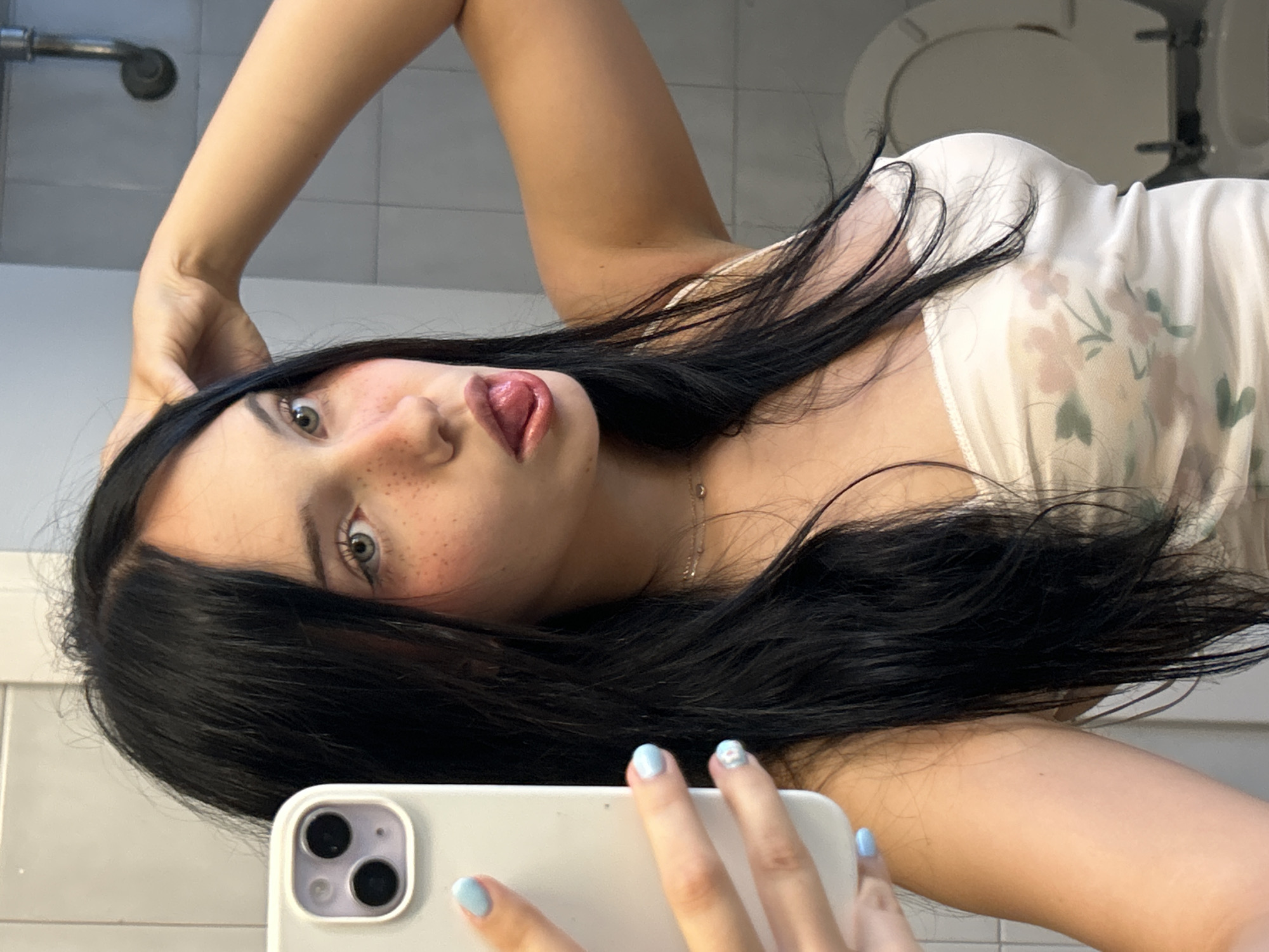 A young woman with long, black hair and fair skin is taking a mirror selfie in a bathroom. She is wearing a white, floral-patterned camisole. Her right hand is raised to her head, while her left hand holds a white smartphone, which is angled to capture her reflection. She is sticking her tongue out and has a playful expression. Her nails are painted a light blue color. The background shows a tiled wall, a sink, and a toilet. The lighting appears to be artificial and originates from above. The image suggests a casual and private setting, likely a personal bathroom. The specific location within San Benedetto del Tronto, Italy cannot be determined from the visual information provided in the image.