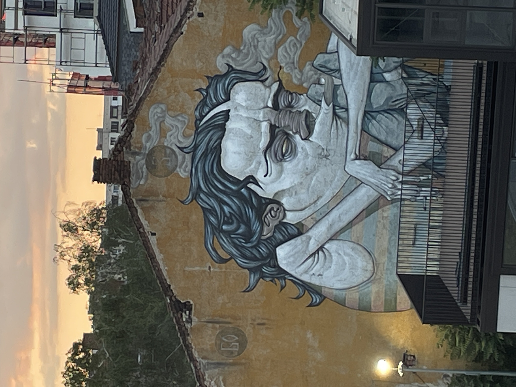 An outdoor shot at golden hour reveals a large-scale mural on the side of an old, damaged building in Sofia, Bulgaria. The mural depicts a stylized, corpulent male figure with dark, wavy hair, exaggerated facial features including a prominent nose, and closed or heavy-lidded eyes. He appears to be leaning on his forearms, wearing a striped garment beneath a light-colored top. Wisps of grey smoke emanate from the right side of his face, suggesting smoking. Above the figure's head on the wall, a circular emblem containing a crescent moon or similar celestial body is surrounded by cloud-like smoke patterns. To the left of the figure, a circular marking on the wall contains the Cyrillic letters "БО". The building facade is a warm yellow-orange, contrasting with the figure's muted blue, grey, and white tones. The building features a terracotta-tiled roof, visibly aged and damaged in places, with a brick chimney stack to the upper right.
In the immediate foreground, a dark metal railing of a balcony or terrace is visible, extending across the lower portion of the image. To the far left, a street light with a bright, illuminated bulb stands beside some lush green foliage.
The background shows an urban landscape bathed in the soft, warm light of either sunrise or sunset. To the left, a dense cluster of trees on an elevated area glows in the light. To the right, modern multi-story buildings, some featuring extensive scaffolding indicating ongoing construction, rise against the sky. Distant residential buildings are also discernible. The sky above is a gradient of soft orange, pink, and light yellow, with subtle cloud formations.