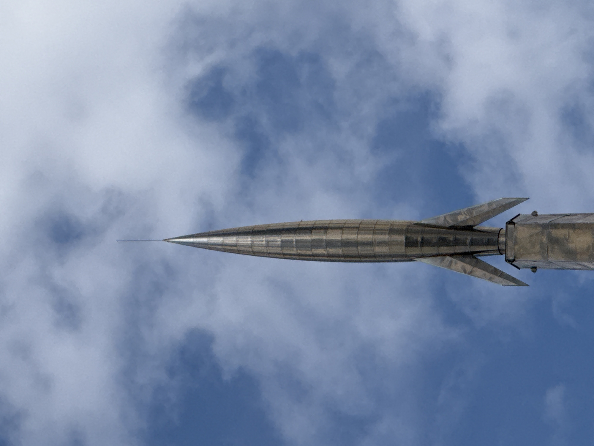 The image shows a close-up, low-angle shot of the Monument "Worker and Kolkhoz Woman" sculpture, located at the All-Russia Exhibition Center (VDNKh) in Moscow, Russia. The sculpture, designed by Vera Mukhina, depicts a dynamic and upward-thrusting male and female figure holding aloft the hammer and sickle. It is made of stainless steel and stands on a pedestal. The sculpture is silhouetted against a bright blue sky filled with white, wispy clouds. The detailed craftsmanship of the sculpture, particularly the textured surface and the sense of movement, is visible. The lighting suggests it is daytime, possibly midday.